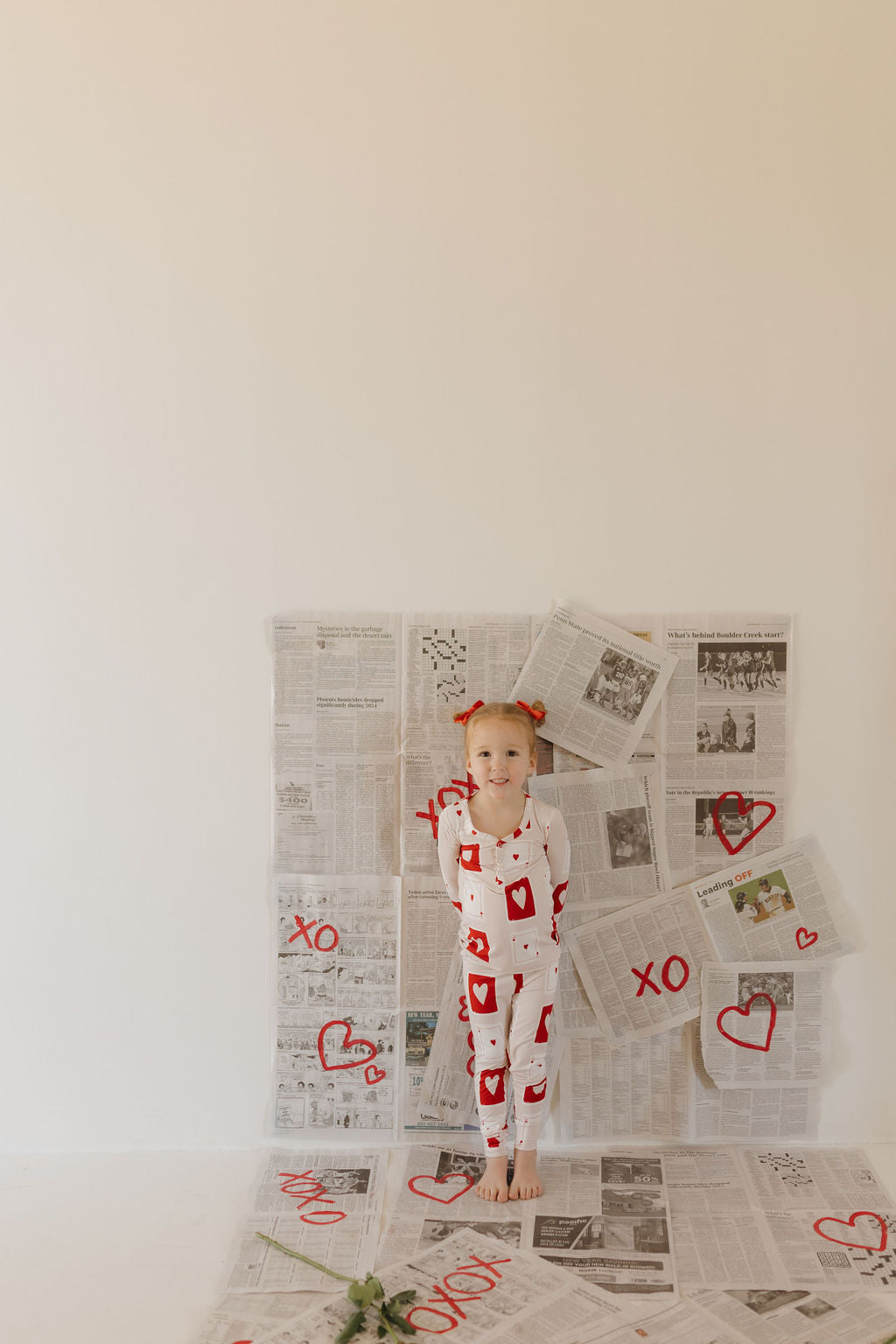 A smiling child in "Love Day" Bamboo Two Piece Pajamas by forever french baby stands barefoot before a newspaper-covered wall adorned with red hearts and "XO". One hand rests on their chin, while a single flower lies on the floor, completing the scene.