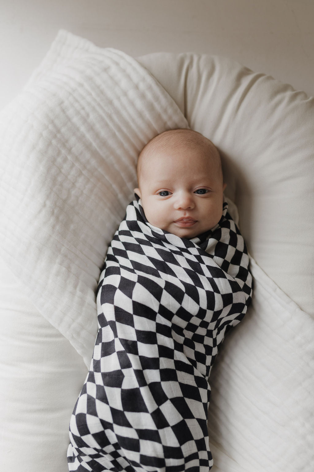 A baby, wrapped in a forever french baby Muslin Swaddle with a black & white wavy checkerboard pattern made from 100% Muslin Cotton, reclines on a soft white pillow. The child's calm gaze is set against light, neutral tones.