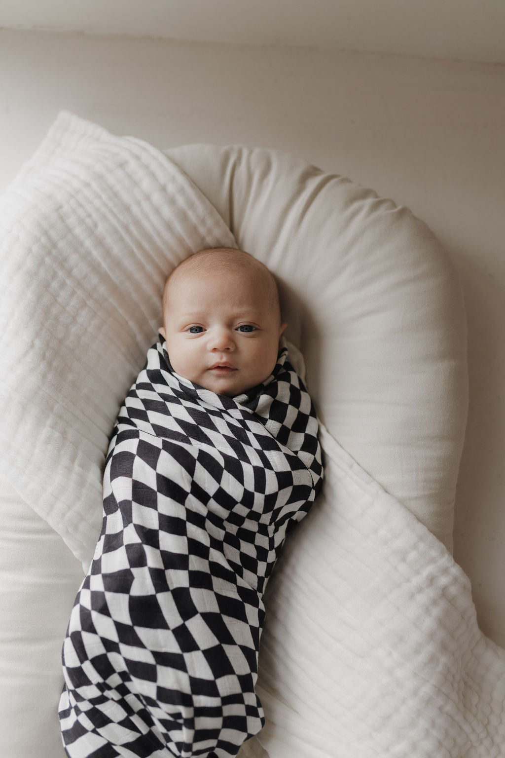 A baby wrapped in a forever french baby Black & White Wavy Checkerboard Muslin Swaddle lies on a white cushioned surface, looking up. The 100% Muslin Cotton swaddle creates a striking contrast with the soft background.