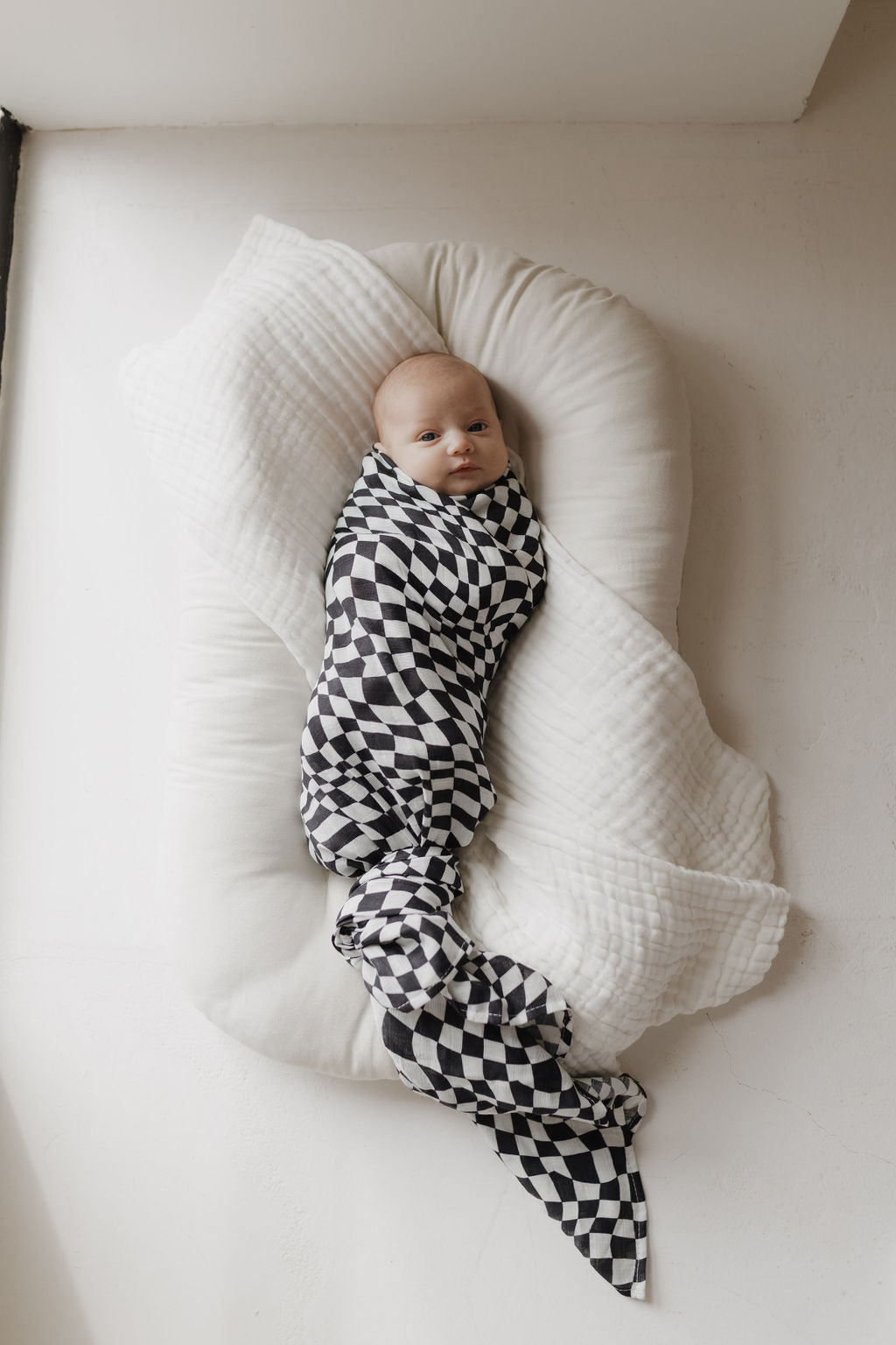 A baby is swaddled in an ultra-soft, black and white wavy checkerboard muslin swaddle by forever french baby, lying on a soft white cushion. The baby looks content in the 100% Muslin Cotton fabric as natural light gently floods the minimalistic setting from the side.