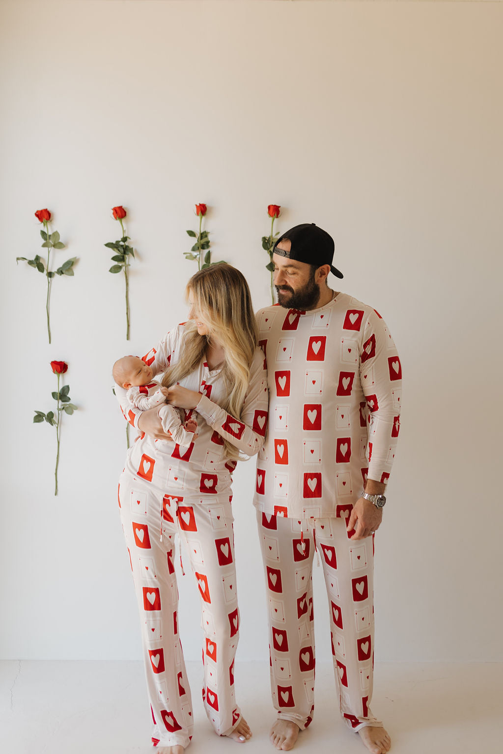 A couple wearing "Love Day" bamboo zip pajamas by forever french baby stands before a rose-adorned wall. The woman holds a baby snug in a blanket, all exuding contentment and connection, while enjoying the cozy comfort of breathable fabric.