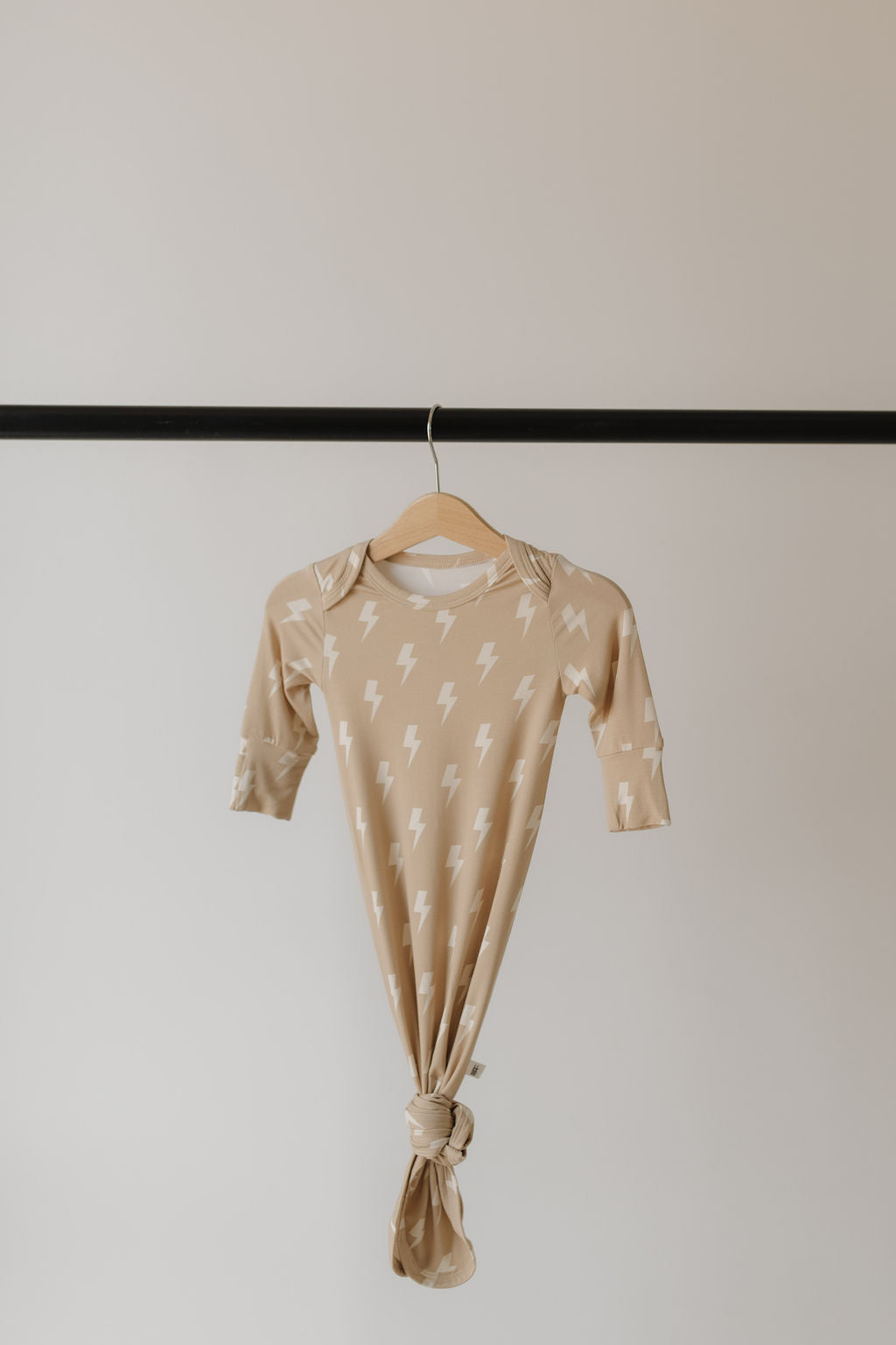 The forever french baby Bamboo Knotted Gown in Tan Lightning Bolt showcases a charming white pattern on hypo-allergenic bamboo, complete with a knotted bottom. It's elegantly displayed on a wooden hanger against a soft, light background. Ideal for newborns!.