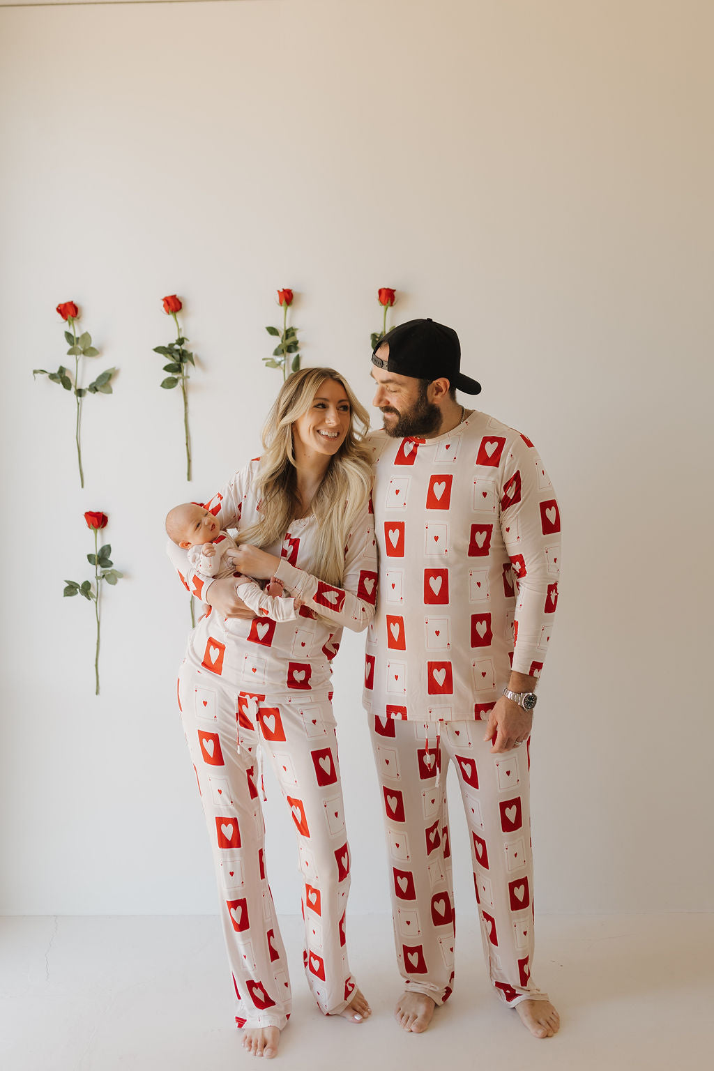 A couple smiles in matching "Love Day" bamboo zip pajamas by forever french baby, holding an infant in the same breathable fabric. Six red roses adorn a light-colored wall behind them.