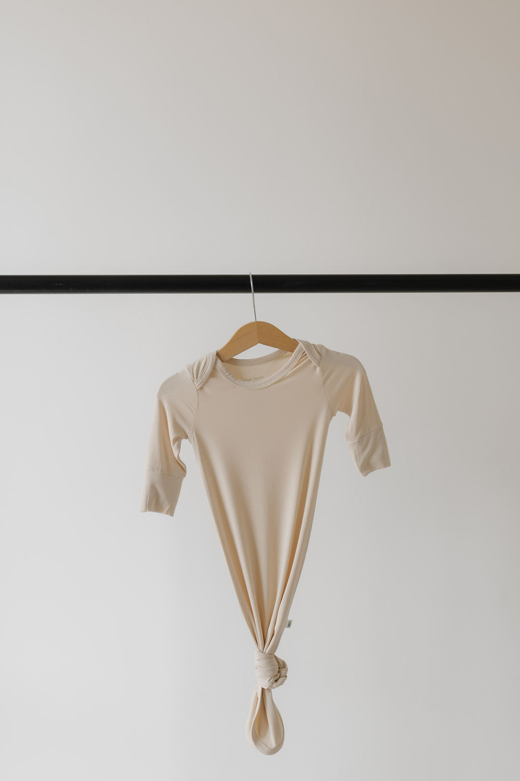 The "Bamboo Knotted Gown | Bone" by forever french baby, crafted from hypo-allergenic bamboo, features long sleeves and a knotted bottom. It hangs elegantly on a wooden hanger displayed on a black rod against a plain white background.