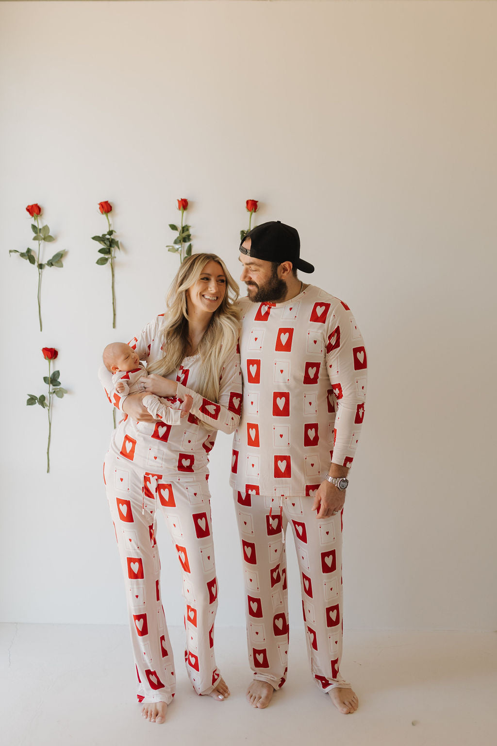 A woman and man, both in forever french baby's Bamboo Zip Pajamas | Love Day with heart patterns, stand together holding a baby. Red roses adorn the white wall behind them. The man wears a cap while the woman smiles warmly at him.