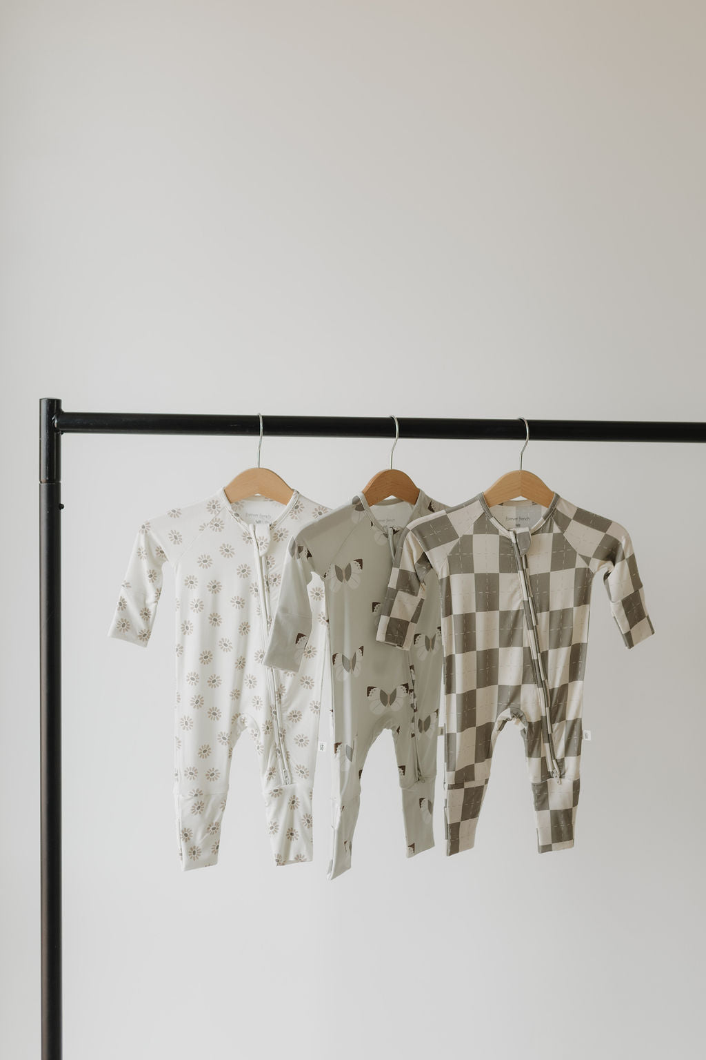 Three hypoallergenic baby onesies by forever french baby, including the Bamboo Zip Pajamas | Daisy Fields, feature floral, abstract animal, and brown & white checkered designs. They hang on wooden hangers from a black metal rack, offering soft comfort for delicate skin.