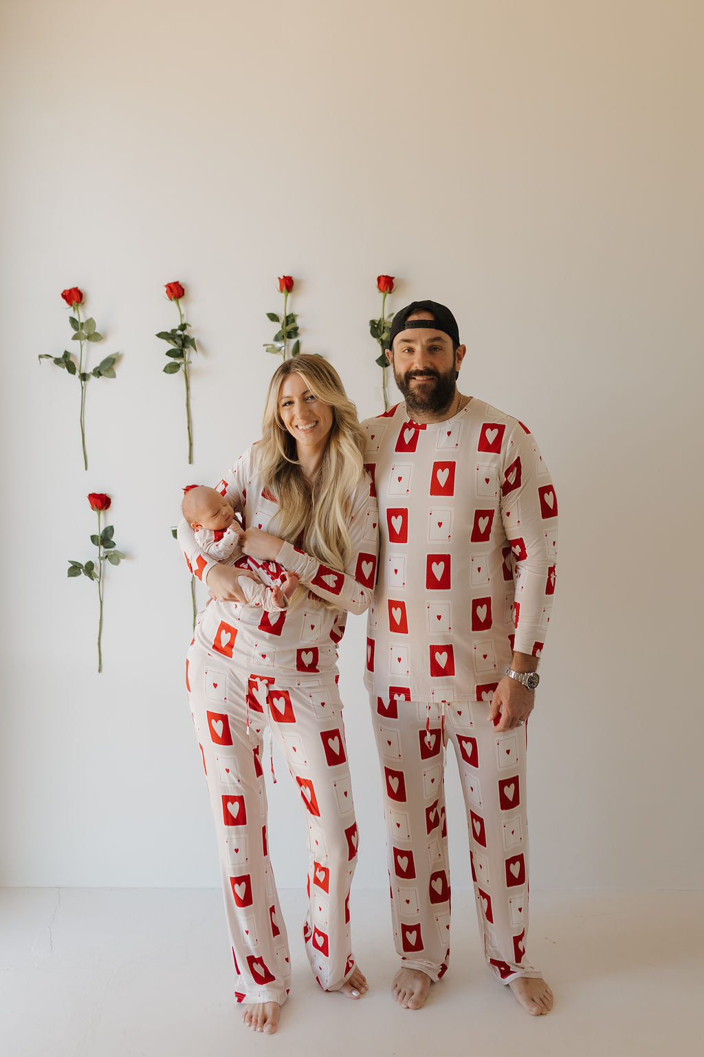 A couple stands barefoot against a plain wall in matching forever french baby Bamboo Zip Pajamas | Love Day, featuring red and white playing card motifs. The woman holds a baby, also in the same cozy pajamas. Five red roses are arranged vertically on the wall behind them.