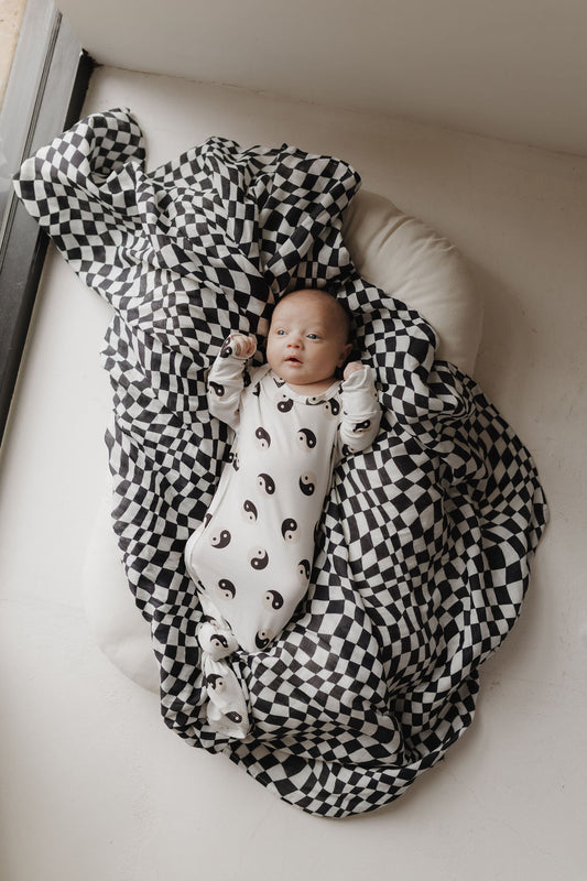 A baby in a white onesie with yin-yang motifs lies on a cushion, wrapped in an ultra-soft "Muslin Swaddle | Black & White Wavy Checkerboard" by forever french baby, creating a cozy and calm scene against a neutral background near a window.