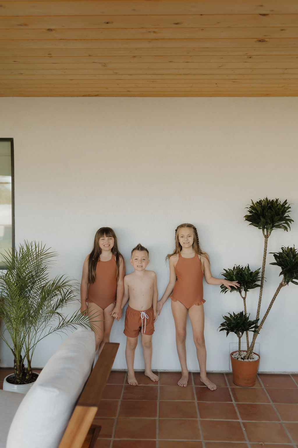 Three children in swimsuits stand on a tiled patio with potted plants. The girls wear matching rust-colored Girls Sleeveless Swimsuit | Terra from forever french baby, while the boy sports rust-colored swim trunks. They smile under a bright outdoor setting with a wooden ceiling above.