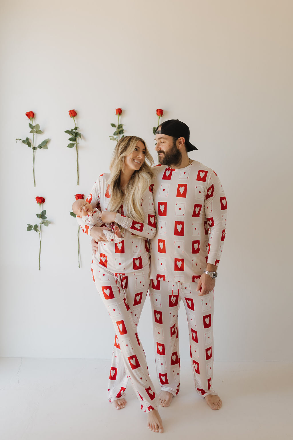 A couple wearing "Bamboo Zip Pajamas | Love Day" by forever french baby, adorned with red hearts, stands with their baby. The man wears a black cap, while six roses are vertically displayed on the bright, simple background. The breathable fabric ensures comfort.