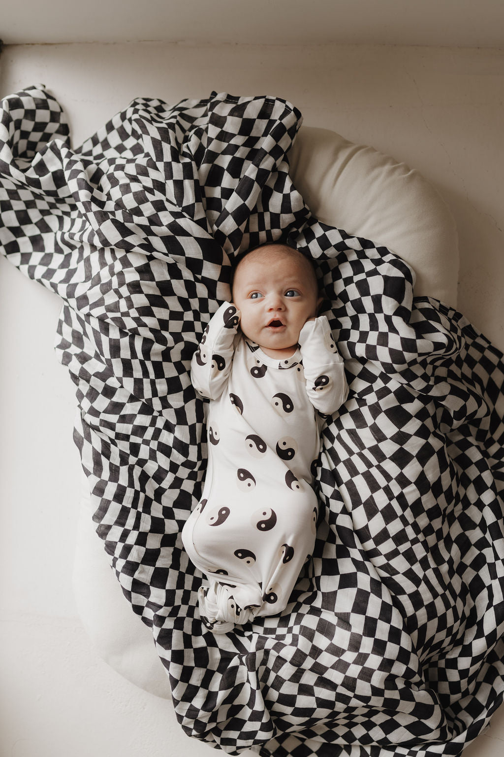 A baby lies on a white cushion, wrapped in an ultra-soft Muslin Swaddle | Black & White Wavy Checkerboard by forever french baby. Wearing a white onesie with yin-yang symbols, the little one gazes upward, hands near their face.