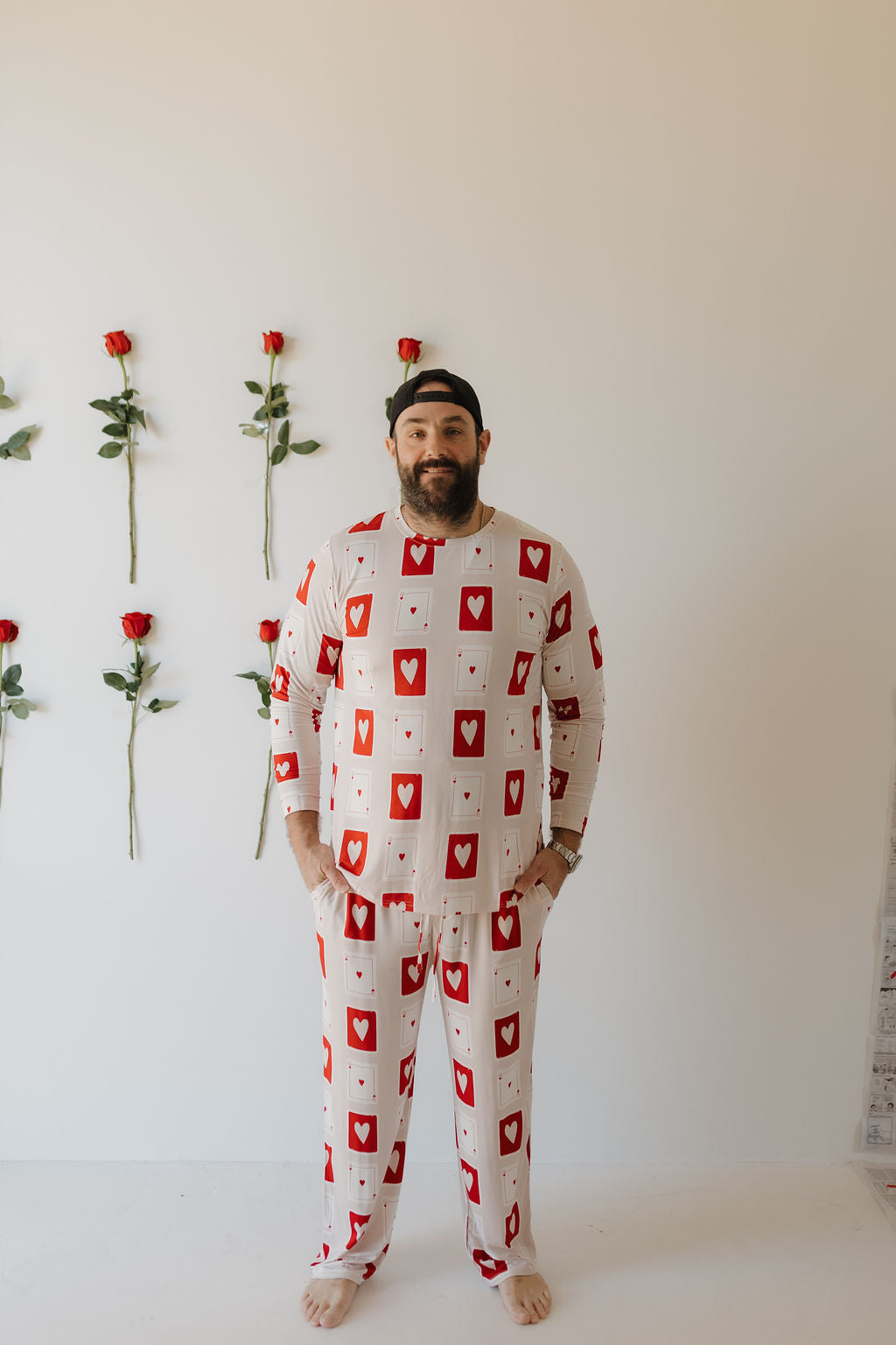 A man wearing the "Men's Bamboo Pajamas | Love Day" by forever french baby, with a playing card design, stands barefoot against a white wall decorated with vertically arranged red roses. He wears a black cap and looks relaxed, appreciating the breathable comfort of his hypo-allergenic pajamas.