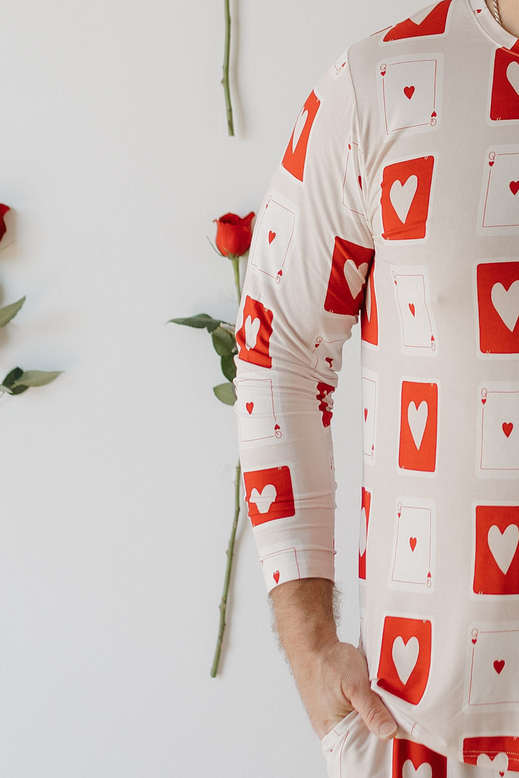 Wearing the forever french baby Men's Bamboo Pajamas | Love Day, featuring a snug, long-sleeved red and white playing card design with hearts, exudes charm. The breathable, hypo-allergenic fabric enhances comfort, while nearby red roses on a white wall create a romantic theme.