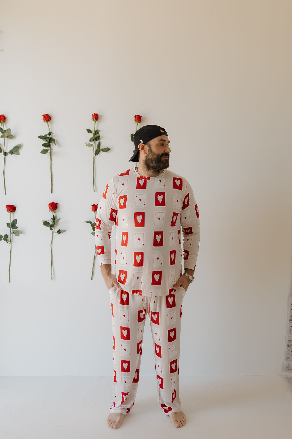 A bearded man stands barefoot against a white wall with vertical rows of red roses, wearing the Love Day pajamas by forever french baby. Adorned with a charming red heart card pattern, he gazes aside, embodying comfort in breathable fabric.