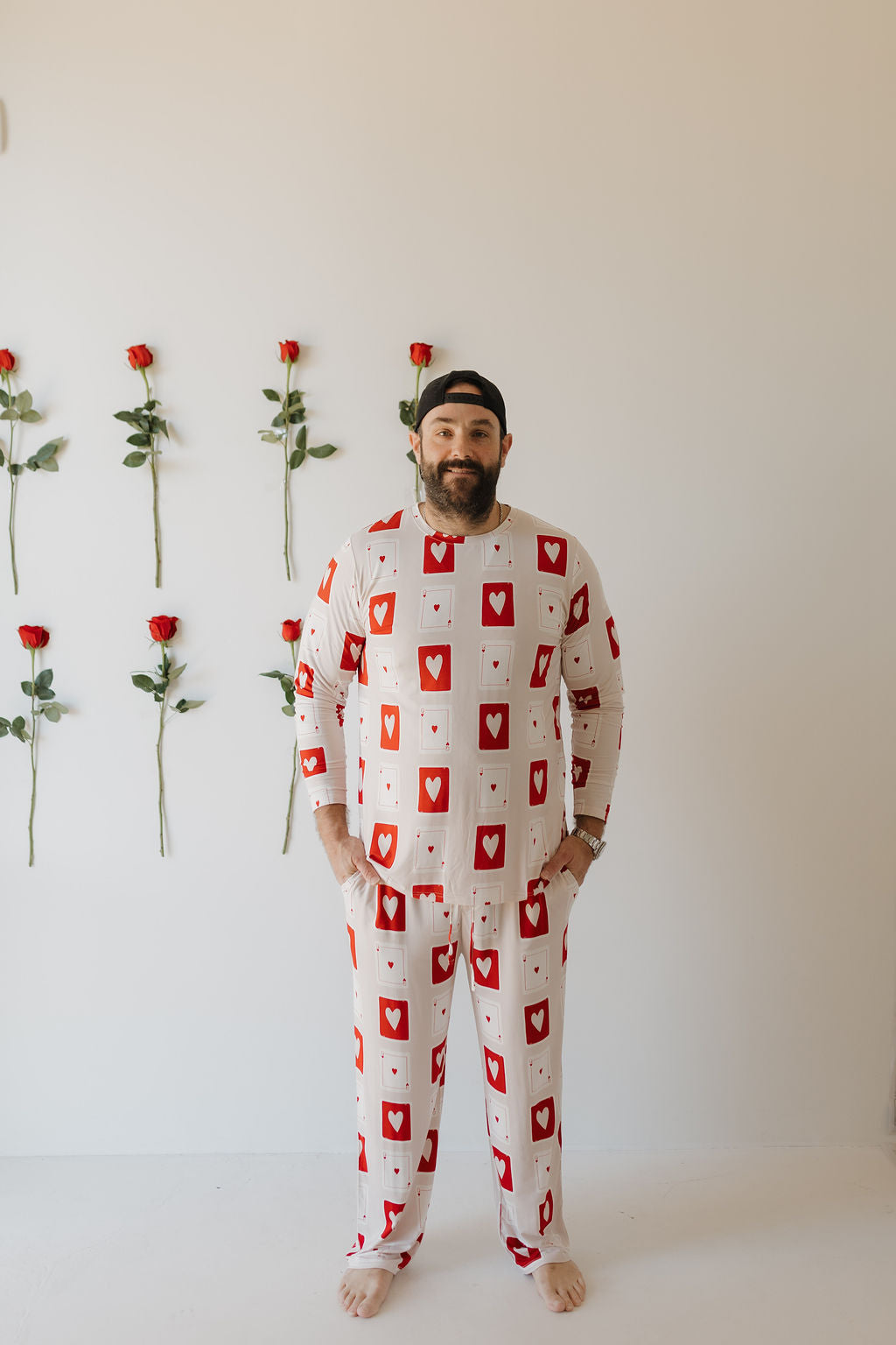 A person stands barefoot against a white wall, comfortably wearing Men's Bamboo Pajamas | Love Day by forever french baby. Adorned with a playing card heart pattern, the hypoallergenic fabric ensures comfort. Five vertical red roses decorate the wall to their left.