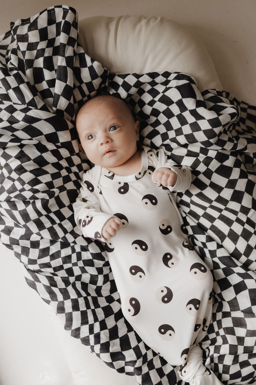 An infant in a white onesie with black yin-yang symbols rests on a forever french baby's ultra-soft Black & White Wavy Checkerboard Muslin Swaddle. Crafted from 100% Muslin Cotton, this blanket enhances the serene scene as the baby gazes upward with bright, calm eyes.