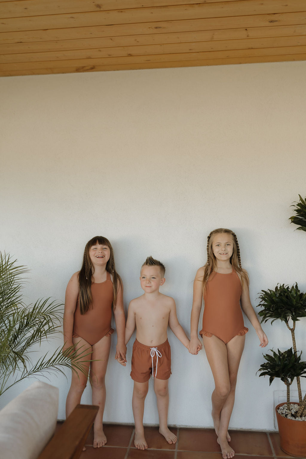 Three children hold hands before a white wall. Two girls wear orange Girls Sleeveless Swimsuits from the Terra collection by forever french baby, flanking a boy in matching swim trunks. Potted plants and a wooden ceiling complete the scene.