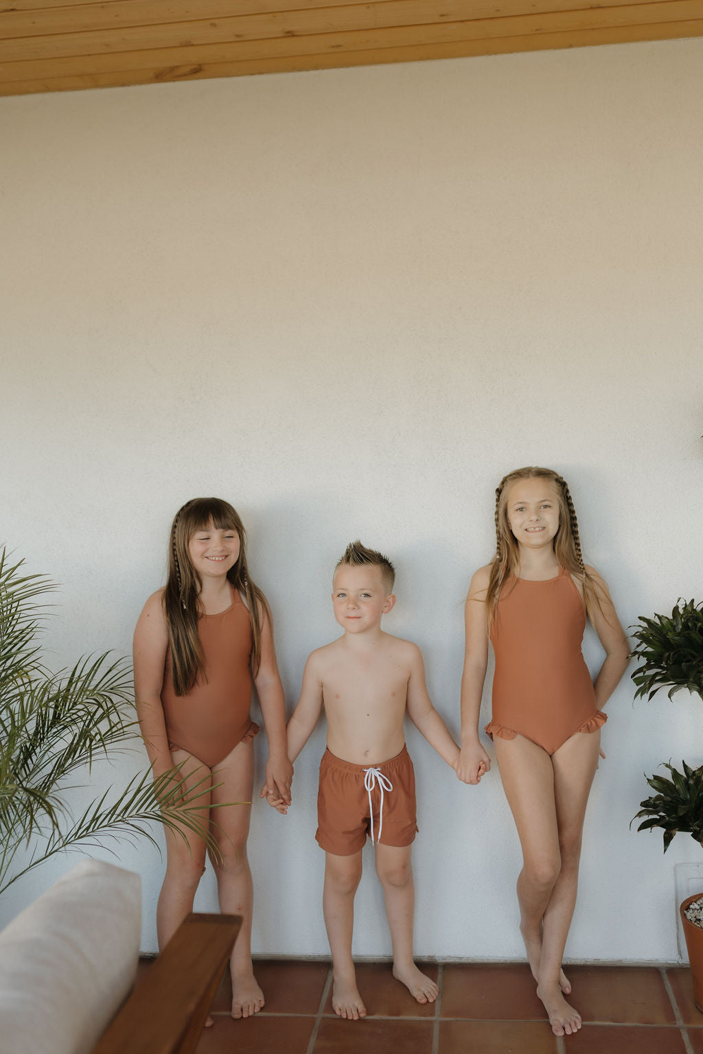 Three children stand holding hands on a tiled floor. Two girls wear matching brown swimsuits from the Terra collection, while the boy in the center sports the "Child Boardshort | Terra" by forever french baby. Potted plants and a wooden ceiling enhance their charming scene.