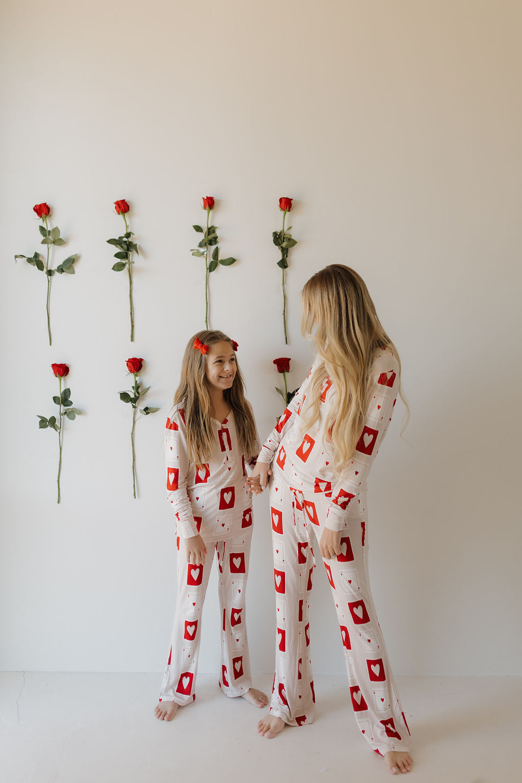 A woman and young girl stand against a white wall with red roses, wearing matching forever french baby Youth Flare Bamboo Pajamas | Love Day. Their hypo-allergenic, breathable fabric with heart and envelope patterns ensures comfort as they share smiles.