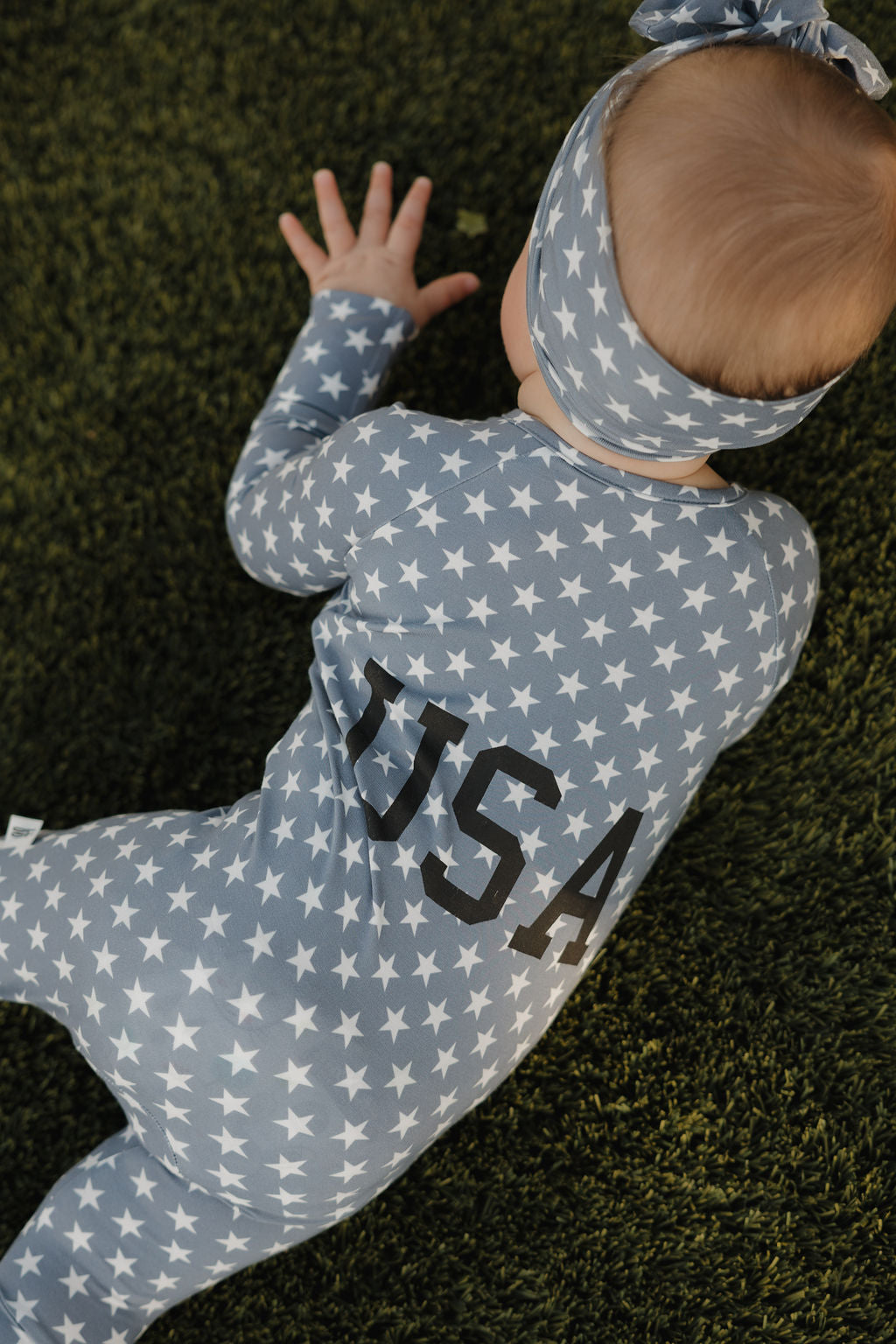 A baby in a blue outfit and the forever french baby Bamboo Headwrap | Star Spangled crawls on green grass.