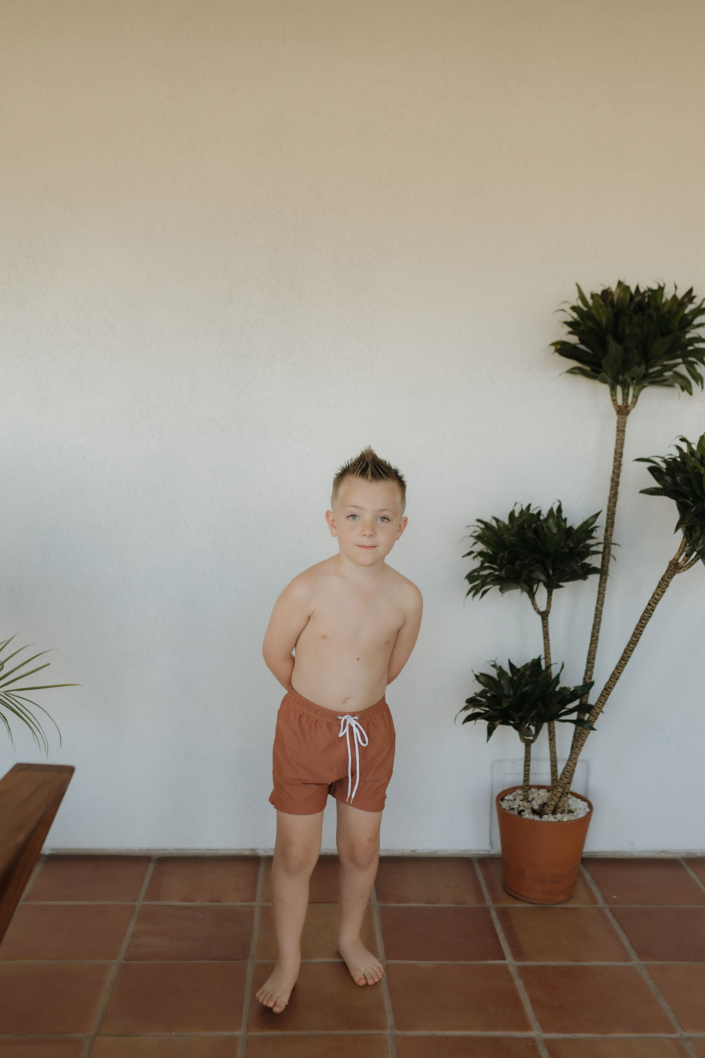 A boy with a spiky haircut stands barefoot on a tiled floor, dressed in "Child Boardshort | Terra" by forever french baby. He poses in front of a white wall flanked by two potted plants, one large and one small, with his hands behind his back.