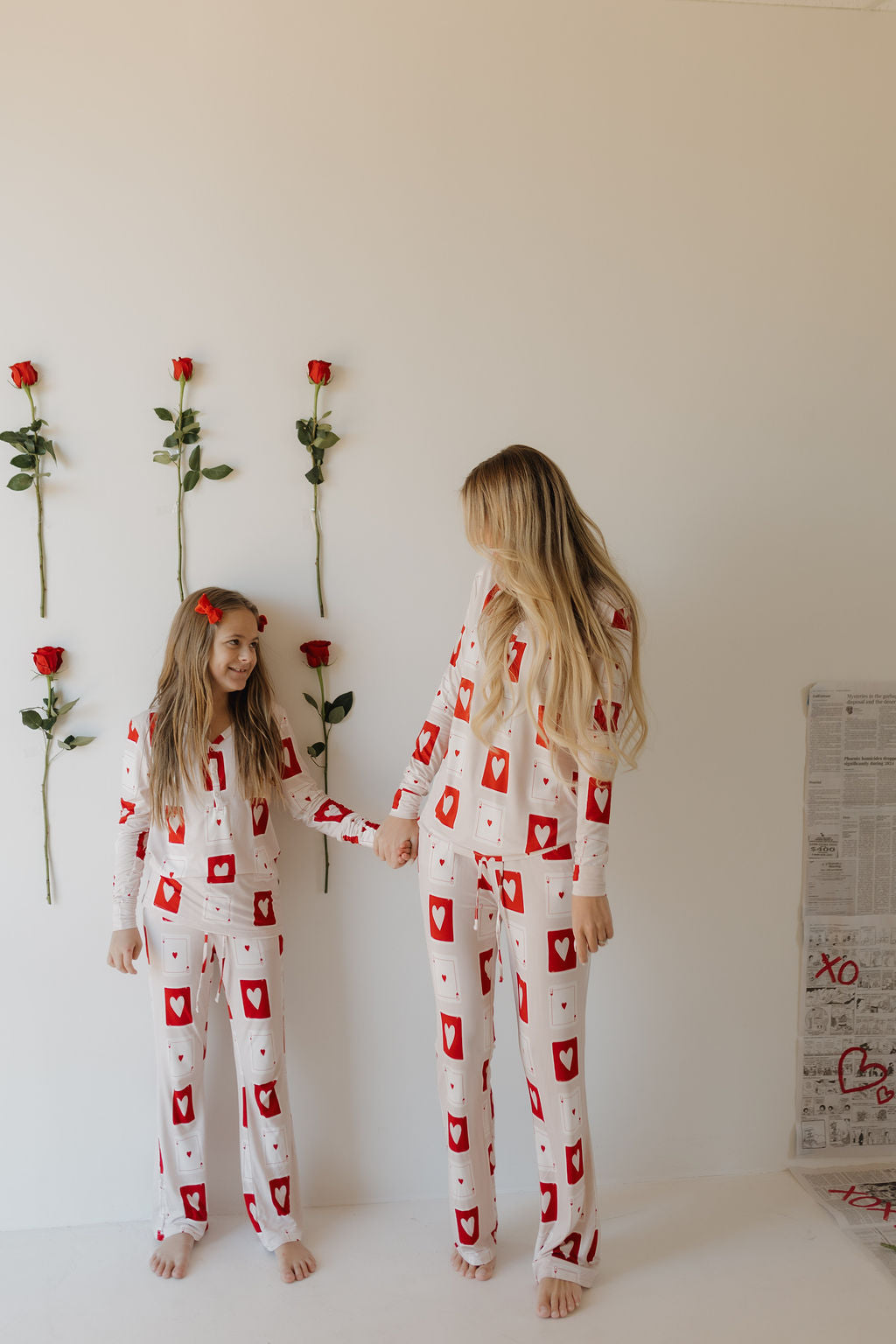 A child and an adult hold hands in matching "Youth Flare Bamboo Pajamas | Love Day" by forever french baby. The white pajamas feature red card suit designs and are made from breathable fabric. They stand before a white wall with red roses while newspapers lie scattered on the floor to the right.