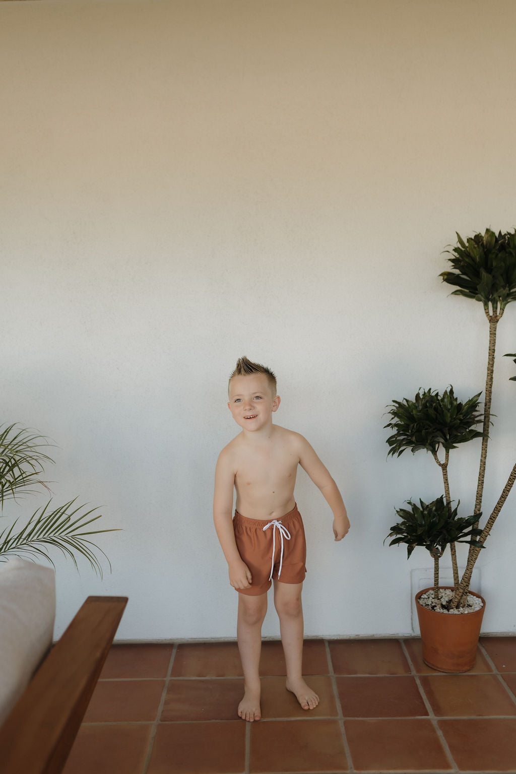 A young child stands on the terracotta floor, wearing forever french baby's Child Boardshort in Terra with a white drawstring for a customizable fit. They smile beneath short hair in the bright room with two potted plants, and part of a wooden seat peeks in from the left.