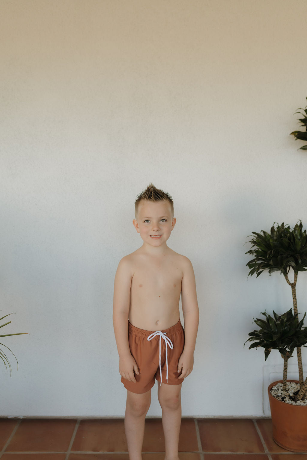 A young boy stands indoors against a white wall, smiling at the camera. Shirtless and wearing forever french baby's Child Boardshort from the Terra collection, he poses with potted plants on either side.