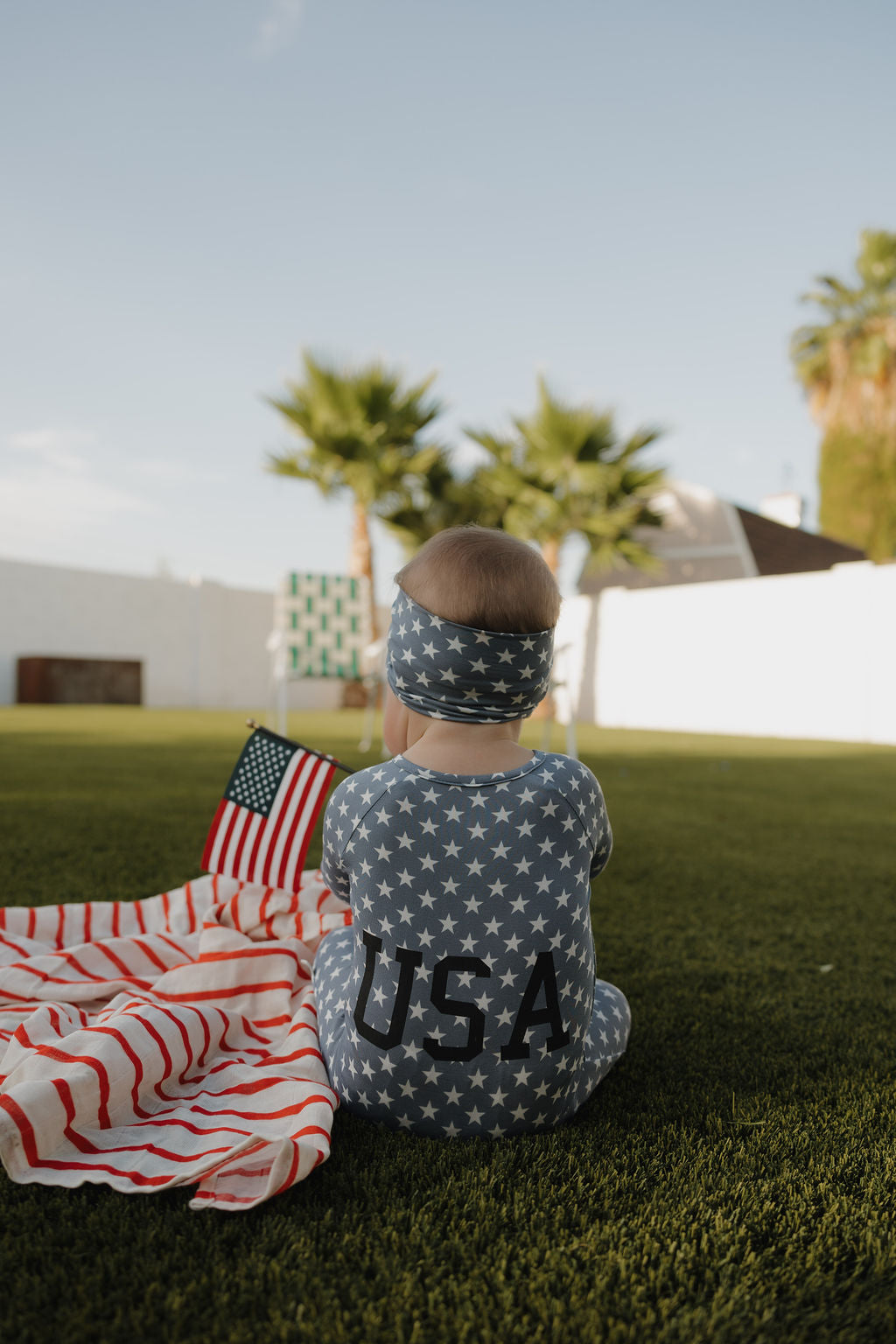 A baby wears the forever french baby Bamboo Headwrap | Star Spangled and a star-patterned "USA" outfit while sitting on grass with an American flag. An American flag blanket, palm trees, and a white fence are in the background.
