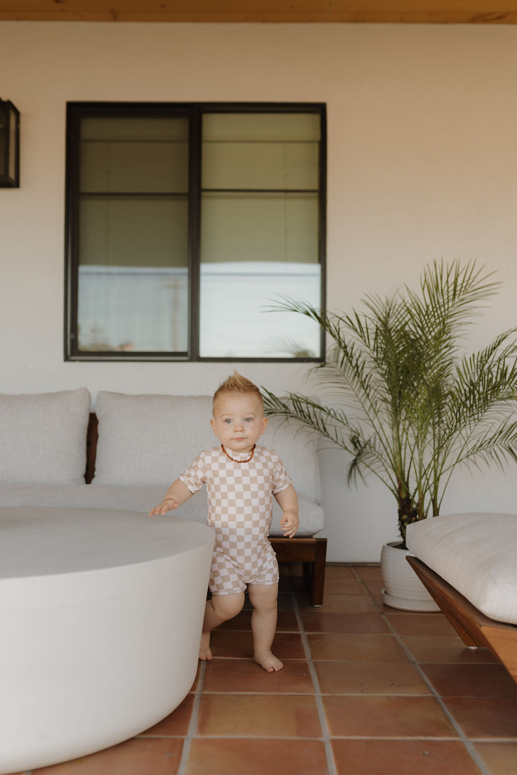 A baby wearing the "Child Shortie Swimsuit | Coastline" from forever french baby stands by a round white table on a tiled patio. A modern sofa with white cushions and a potted palm add charm, while a rectangular window adorns the wall.