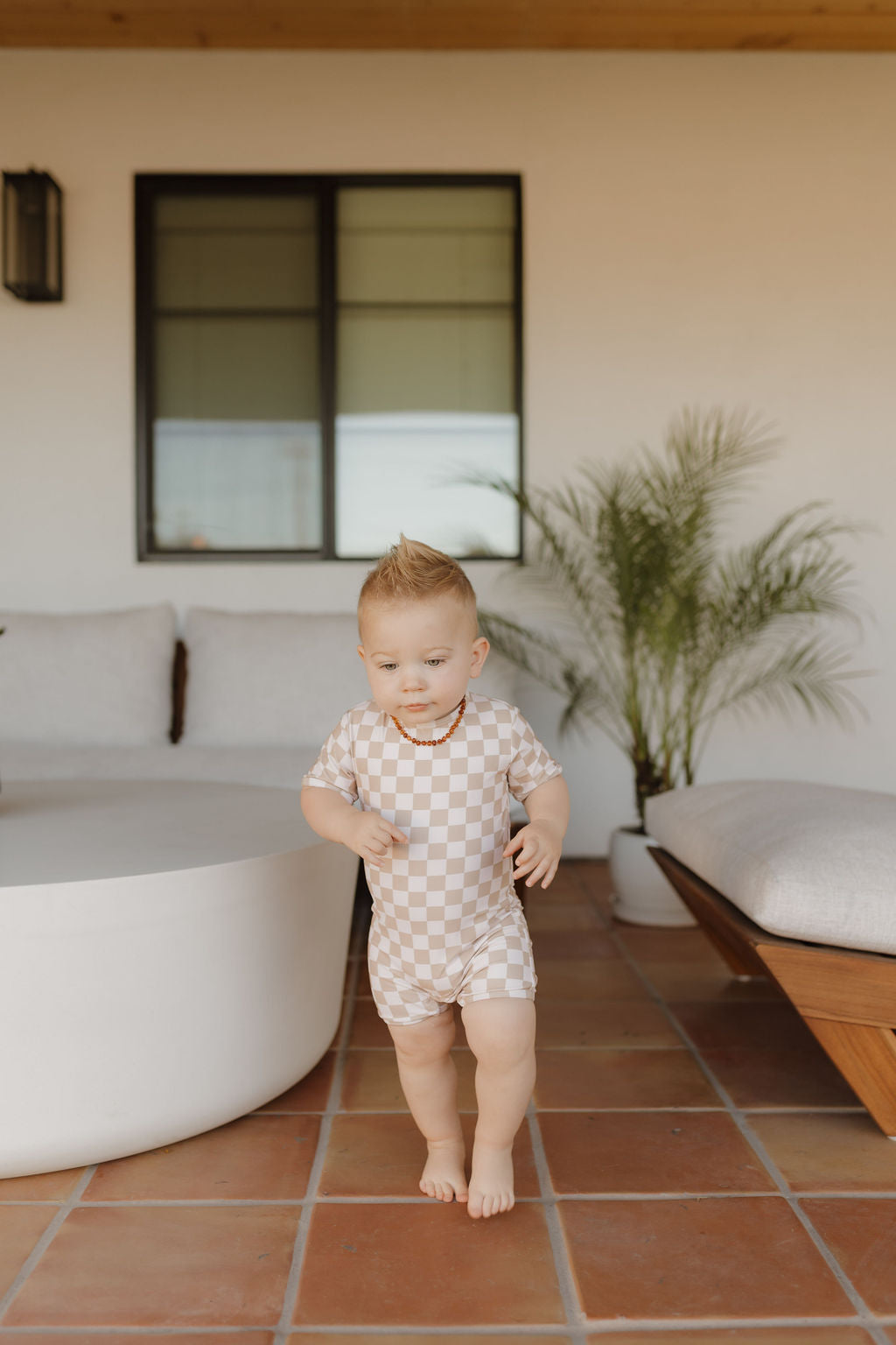 A toddler in a light brown checkered Child Shortie Swimsuit from the Coastline Collection by forever french baby stands on a tiled patio, stepping forward. A round white table, cushioned bench, potted plant, and window create a warm, cozy scene.