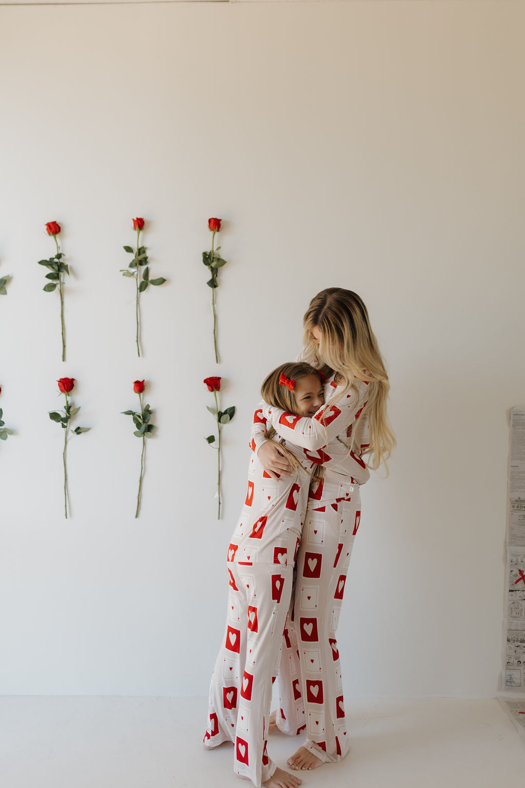 A woman and young girl hug in Youth Flare Bamboo Pajamas from forever french baby, featuring heart patterns. They stand in a bright room with single red roses lined up on the wall.