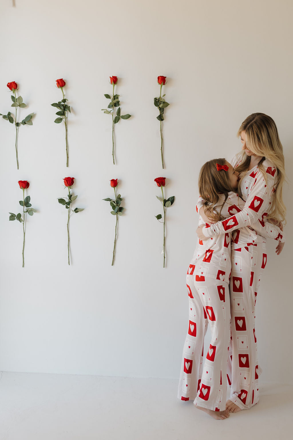 A woman and child in matching hypo-allergenic pajamas from "forever french baby," featuring the Youth Flare Bamboo Pajamas | Love Day with white fabric and red heart designs, hug in front of a white wall graced by nine red roses arranged in three rows.