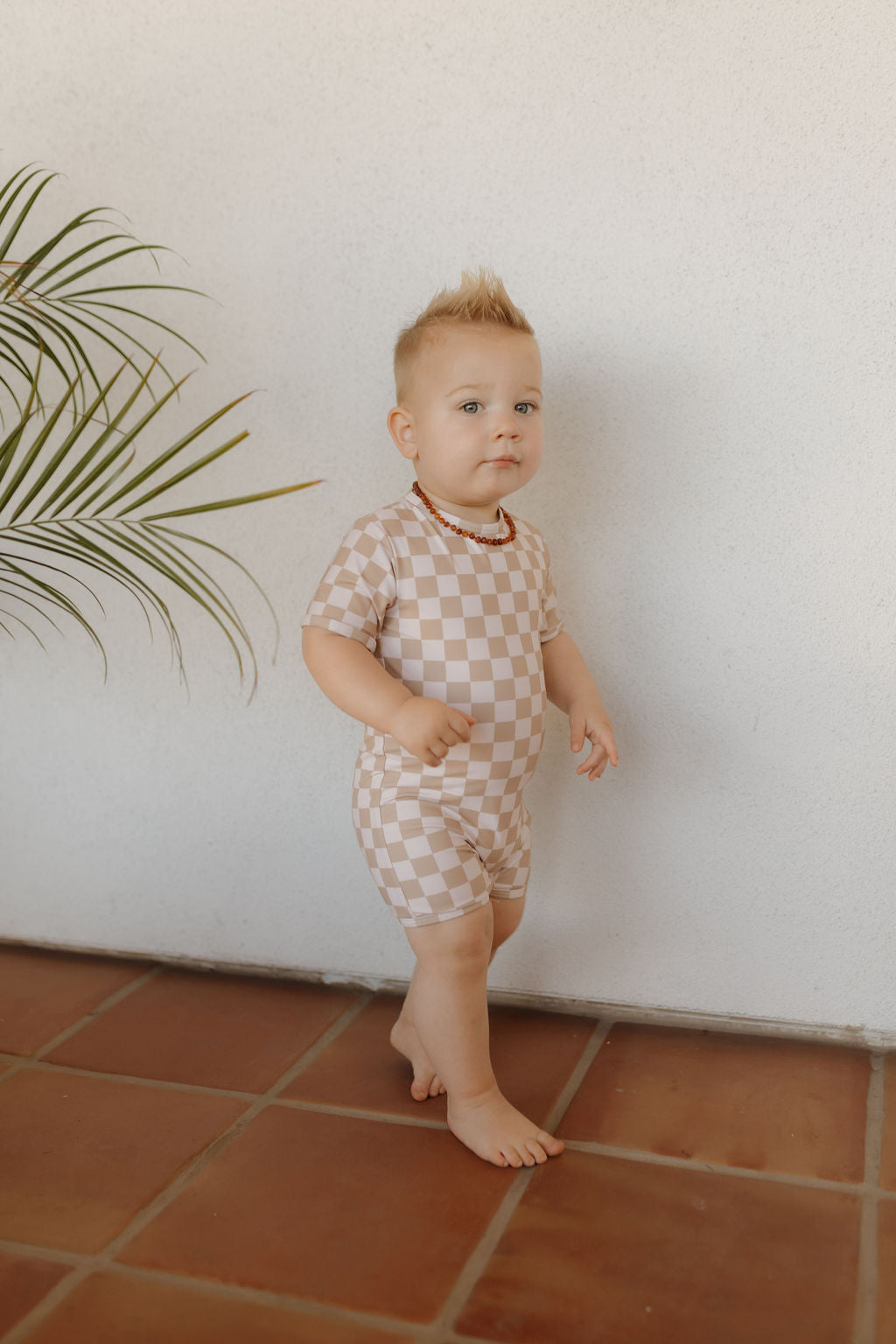 A toddler with a sandy blonde mohawk, barefoot on a tiled floor, flaunts the breezy style of the Coastline Collection in a beige and white checkered romper with a beaded necklace. A palm leaf partially appears on the left against a light wall. Product: Child Shortie Swimsuit | Coastline by forever french baby.