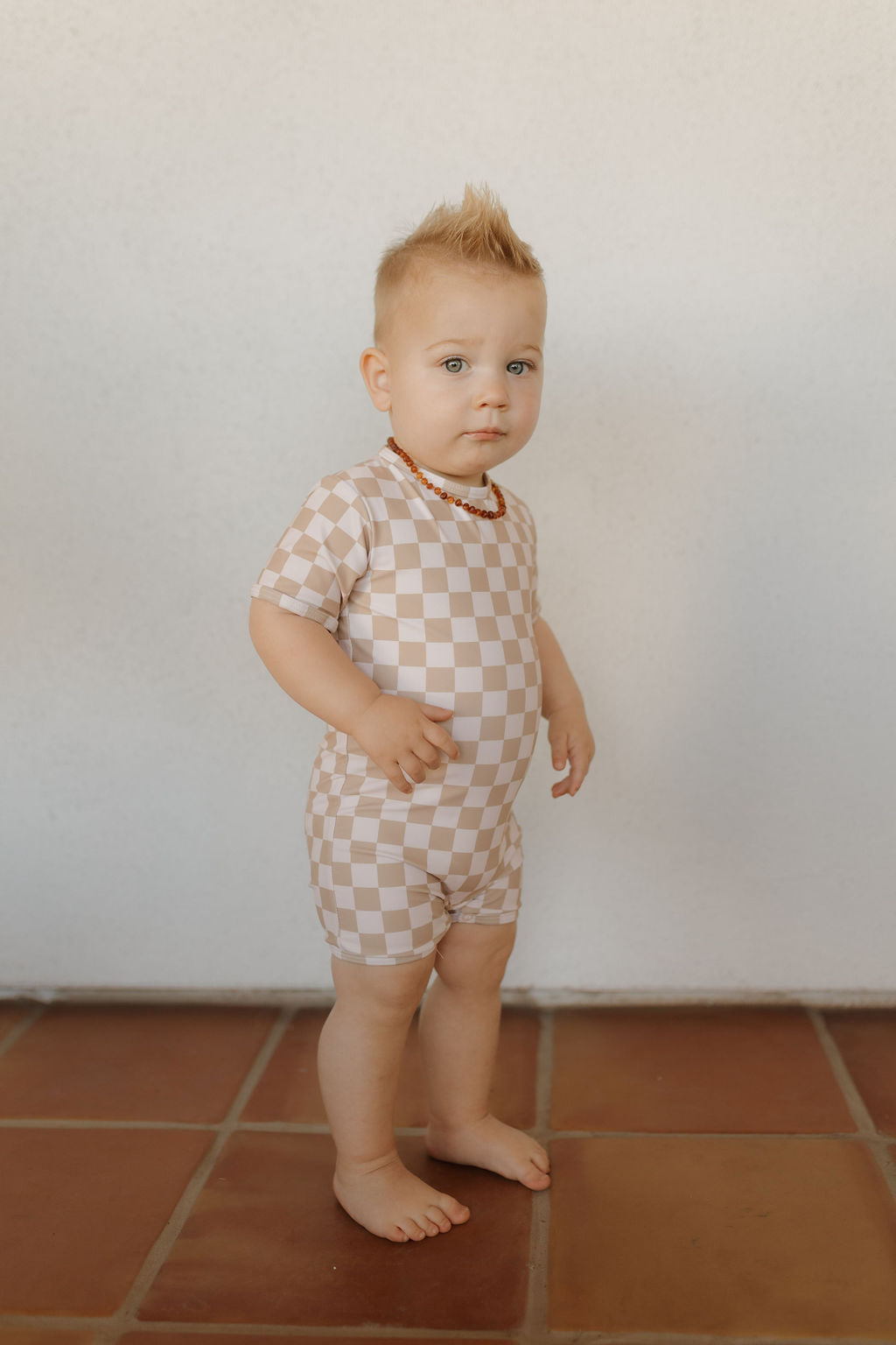 A toddler on a tiled floor wears the Child Shortie Swimsuit from forever french baby's Coastline Collection. With a short mohawk and beaded necklace, the child poses against an off-white wall backdrop.