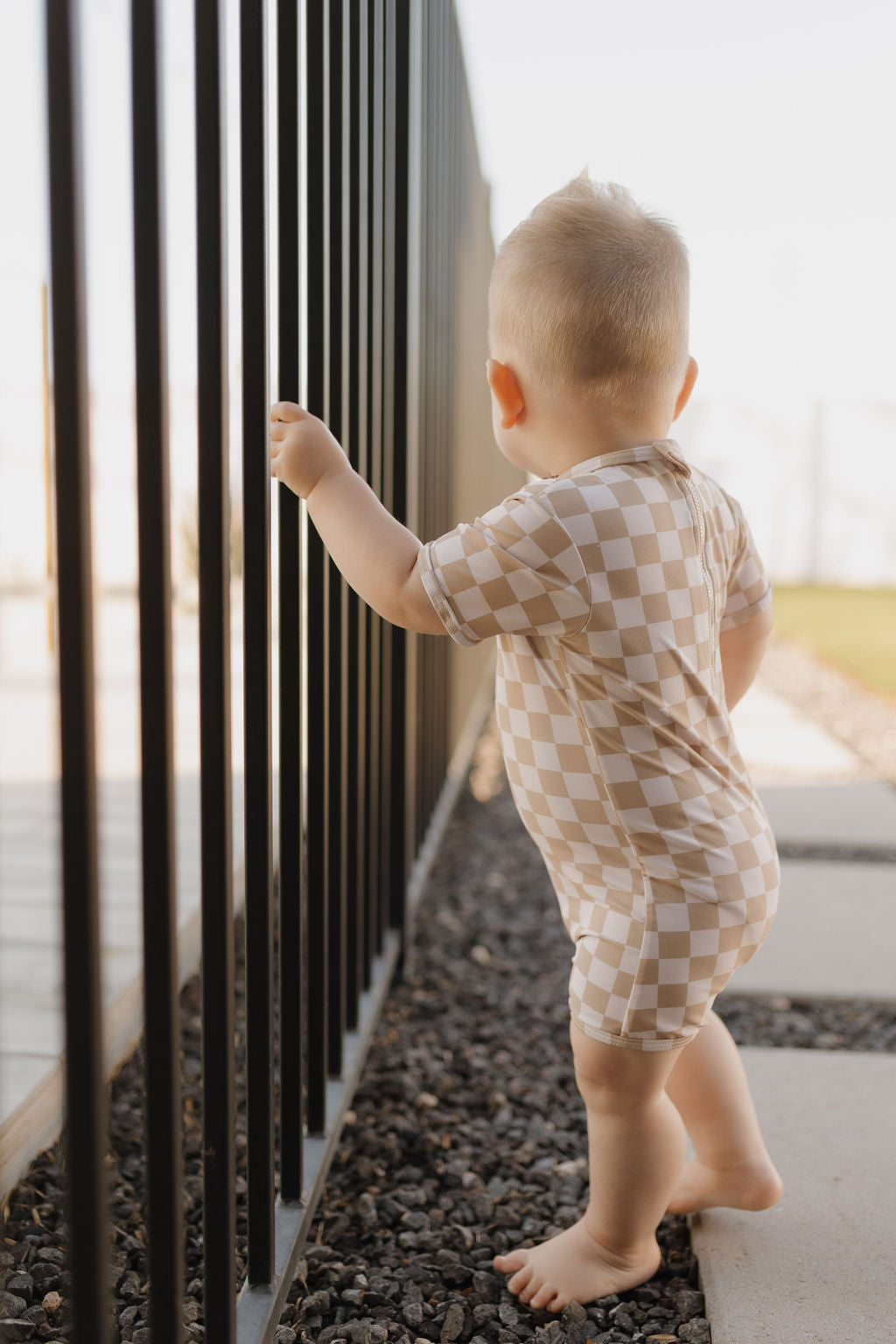 In a sunny setting, a toddler in the "Child Shortie Swimsuit" from forever french baby's Coastline Collection stands on a pebble path, gripping a black metal fence.