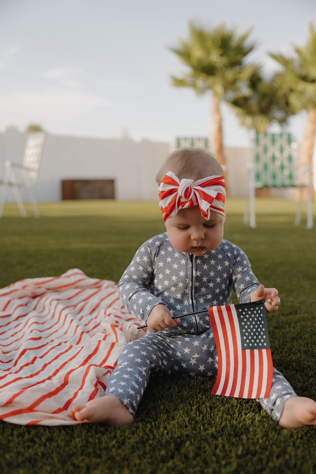 A baby in forever french baby's Bamboo Zip Pajamas | Star Spangled sits on grass, wearing a red, white, and blue headband and holding an American flag beside a striped blanket. Palm trees and outdoor chairs are in the background.