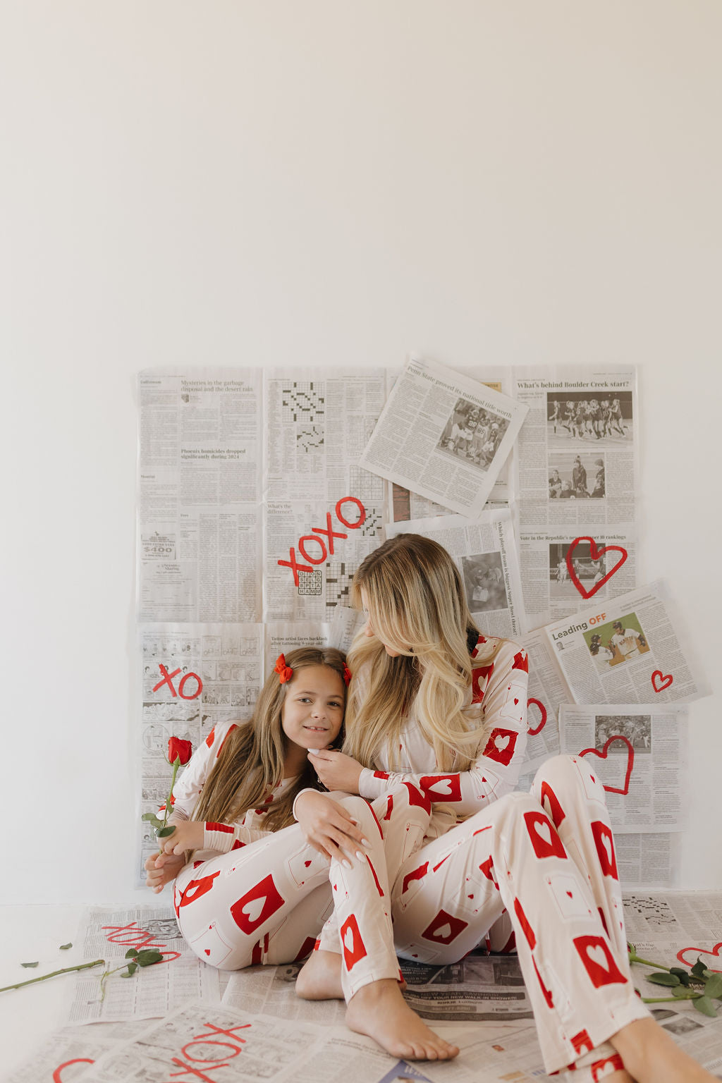 A woman and girl share an affectionate moment in matching "Youth Flare Bamboo Pajamas | Love Day" by forever french baby, surrounded by heart-adorned newspaper and roses. The hypo-allergenic, breathable fabric enhances their comfort.