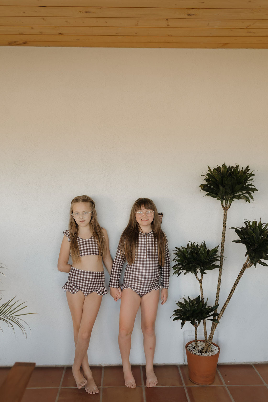 Two young girls in Chocolate Gingham swimwear from forever french baby stand barefoot on a tiled floor next to a potted plant, posing against a pale wall under a wooden ceiling.