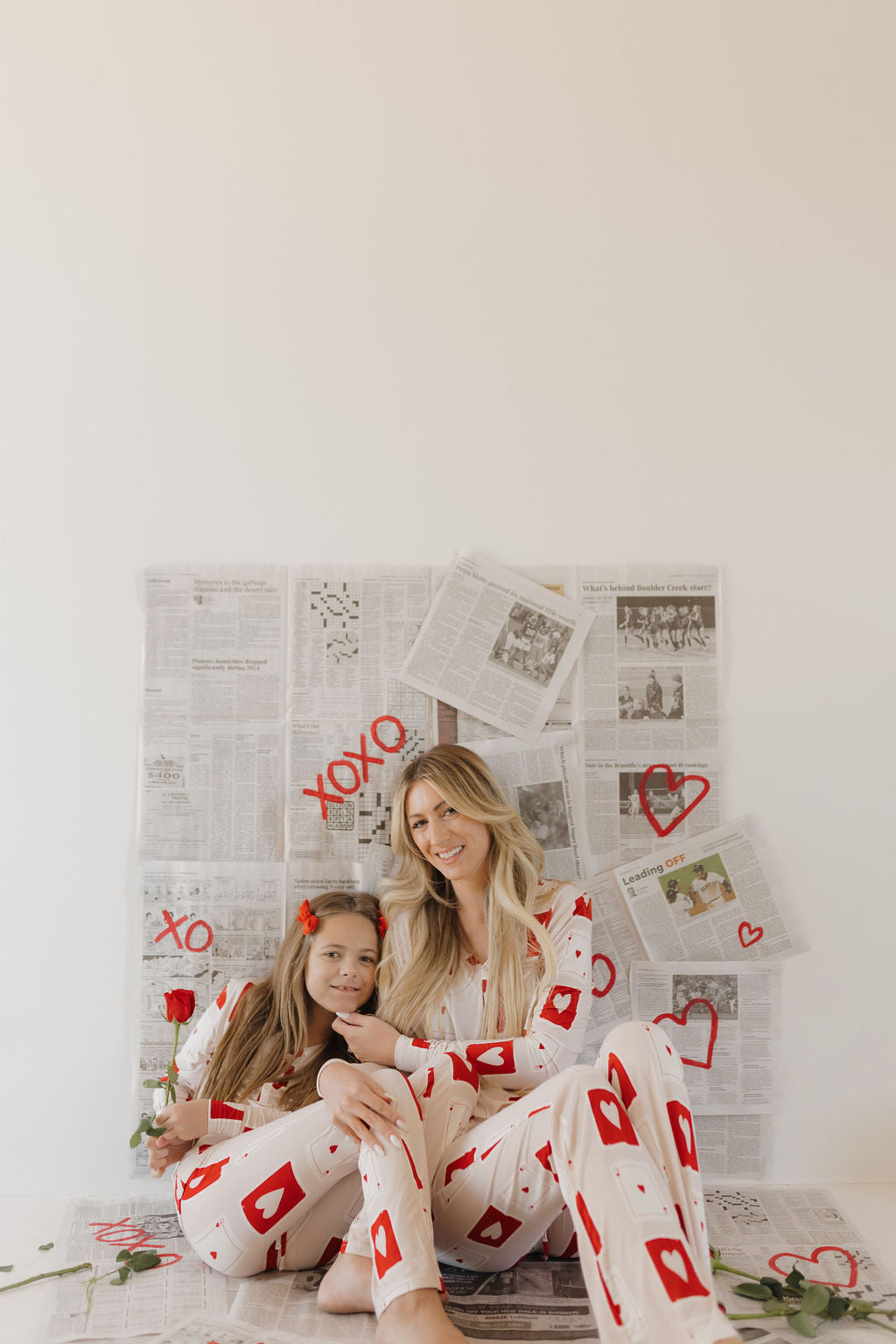 A woman and a girl sit on the floor in their Youth Flare Bamboo Pajamas | Love Day from forever french baby, featuring red heart designs. They smile at the camera with a backdrop of newspaper and heart decorations, while the girl holds a red rose.