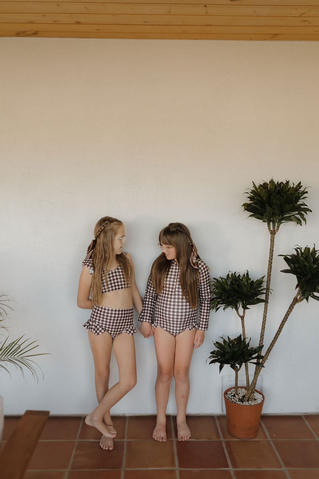 Two girls stand on a tiled patio in matching Girls Two Piece Swimsuits from the Chocolate Gingham collection by forever french baby, gazing at each other with a potted plant nearby, capturing timeless charm and style.