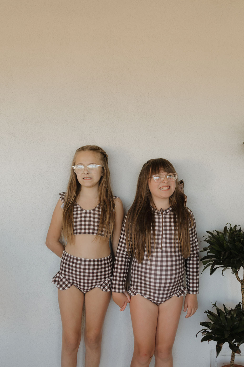 Two young girls, sporting long hair and protective glasses, stand hand in hand against a plain background. They're wearing the "Girls Two Piece Swimsuit | Chocolate Gingham" from forever french baby. A small plant on the side adds a natural touch to this charming scene.