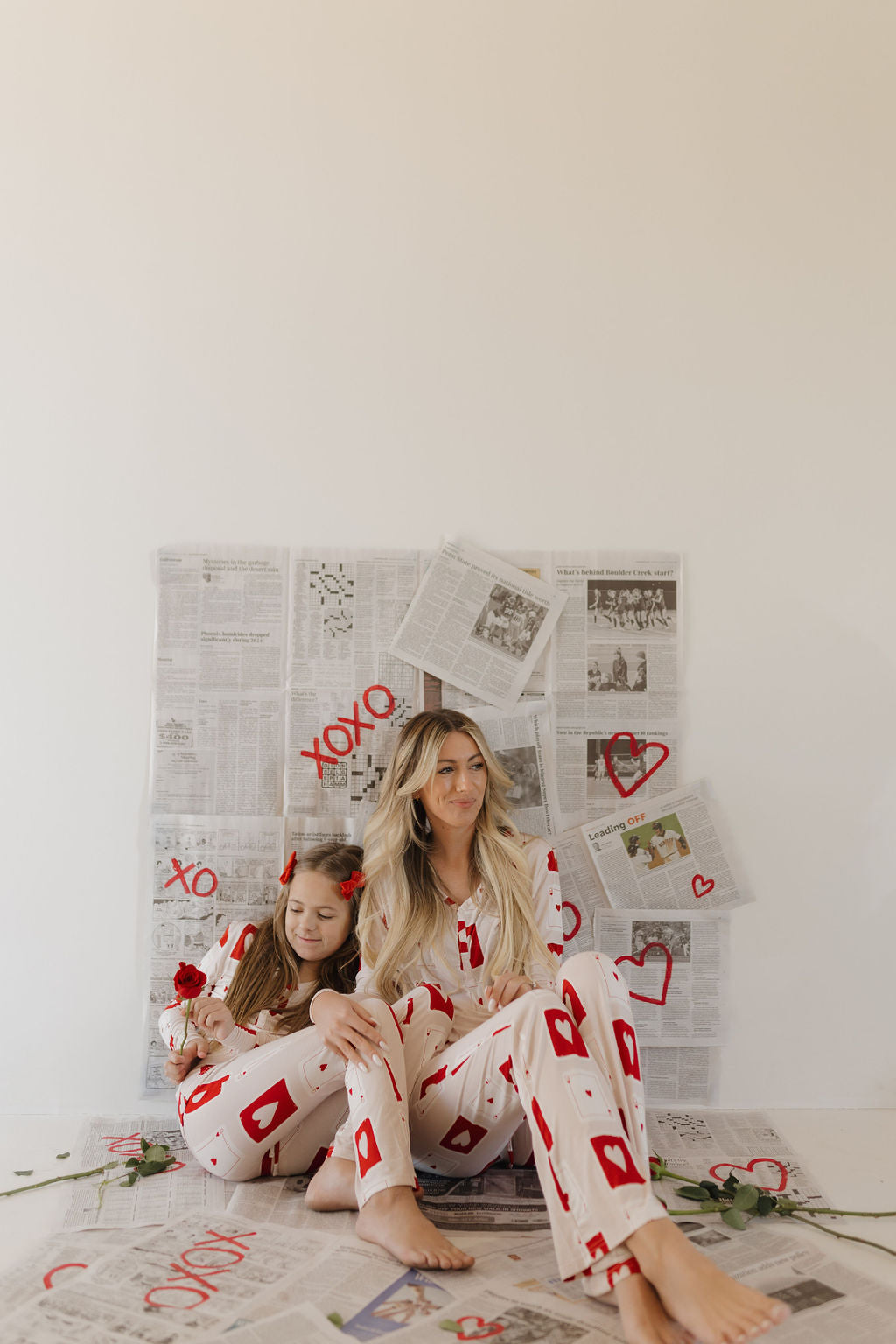 A woman and child sit on the floor in matching forever french baby Youth Flare Bamboo Pajamas | Love Day, featuring red card suits. Surrounding them are newspaper pages with red Xs, Os, and hearts. The child holds a rose as the breathable fabric embraces them for comfort.