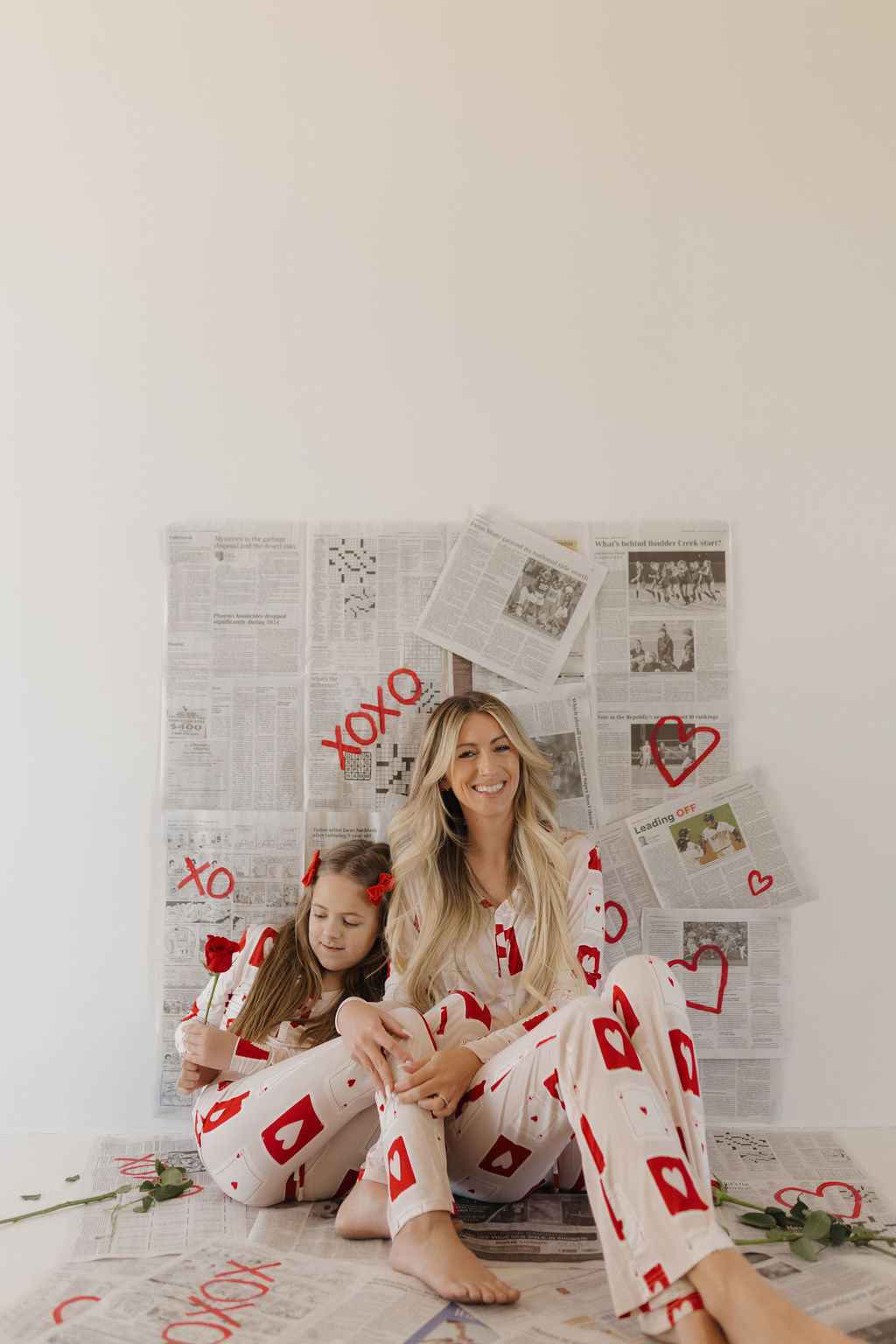 A smiling woman and a young girl wearing forever french baby's Youth Flare Bamboo Pajamas | Love Day, sit on a newspaper-covered floor. The comfortable bamboo fabric features red card suits as the girl holds a rose against an "XOXO"-adorned backdrop.