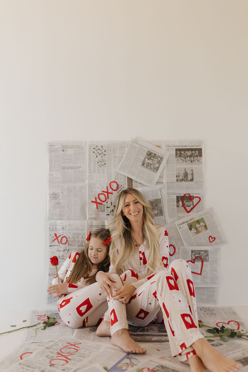 A woman and a young girl sit on the floor against a newspaper-covered wall, wearing matching hypo-allergenic "Love Day" pajamas by forever french baby. The girl holds a red rose, and red hearts with "XOXO" are drawn on the newspapers.