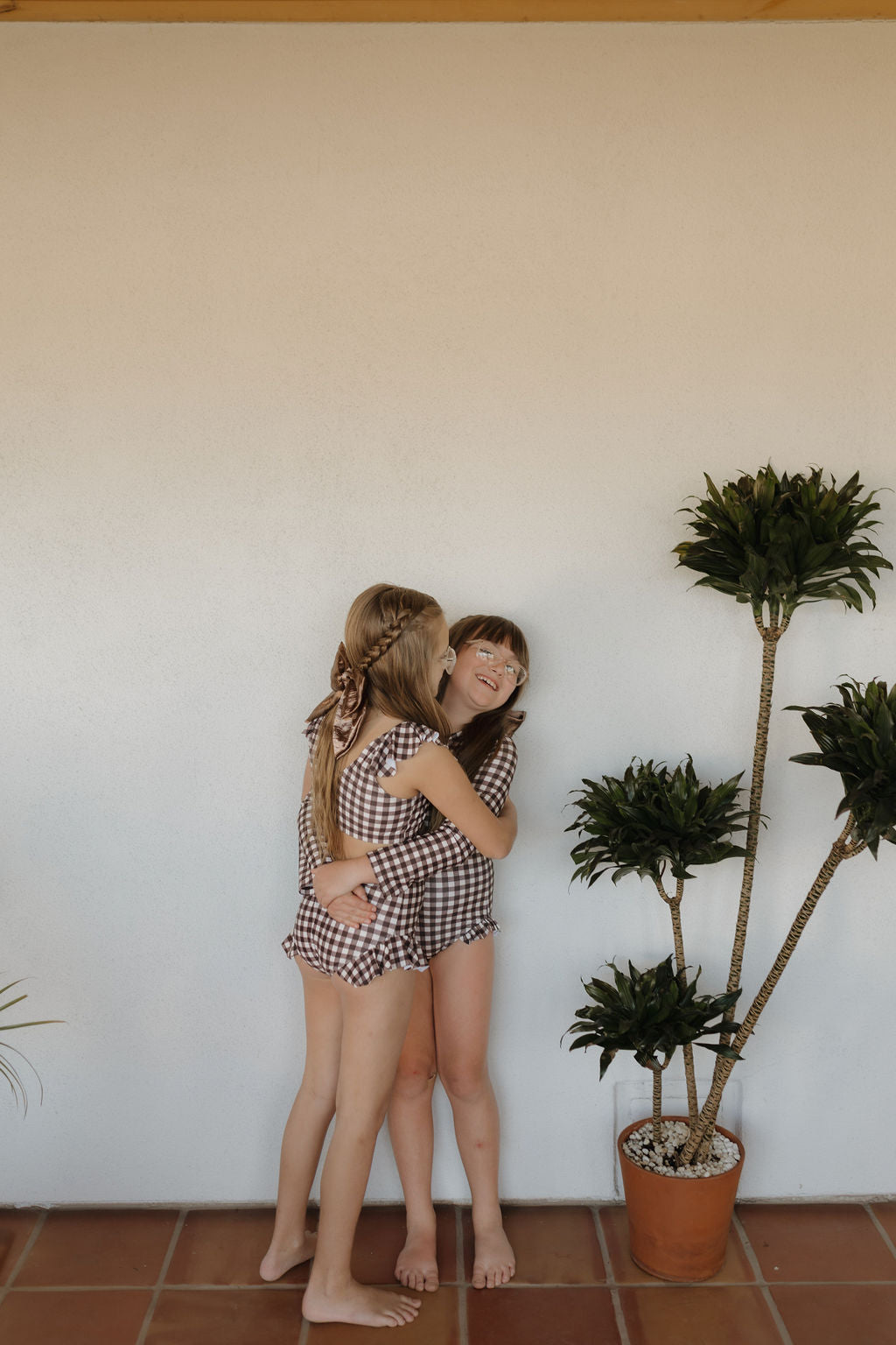 Two young girls in a Chocolate Gingham Girls Two Piece Swimsuit by forever french baby embrace and smile, standing barefoot on terracotta tiles next to a tall potted plant against a white wall, embodying timeless charm.