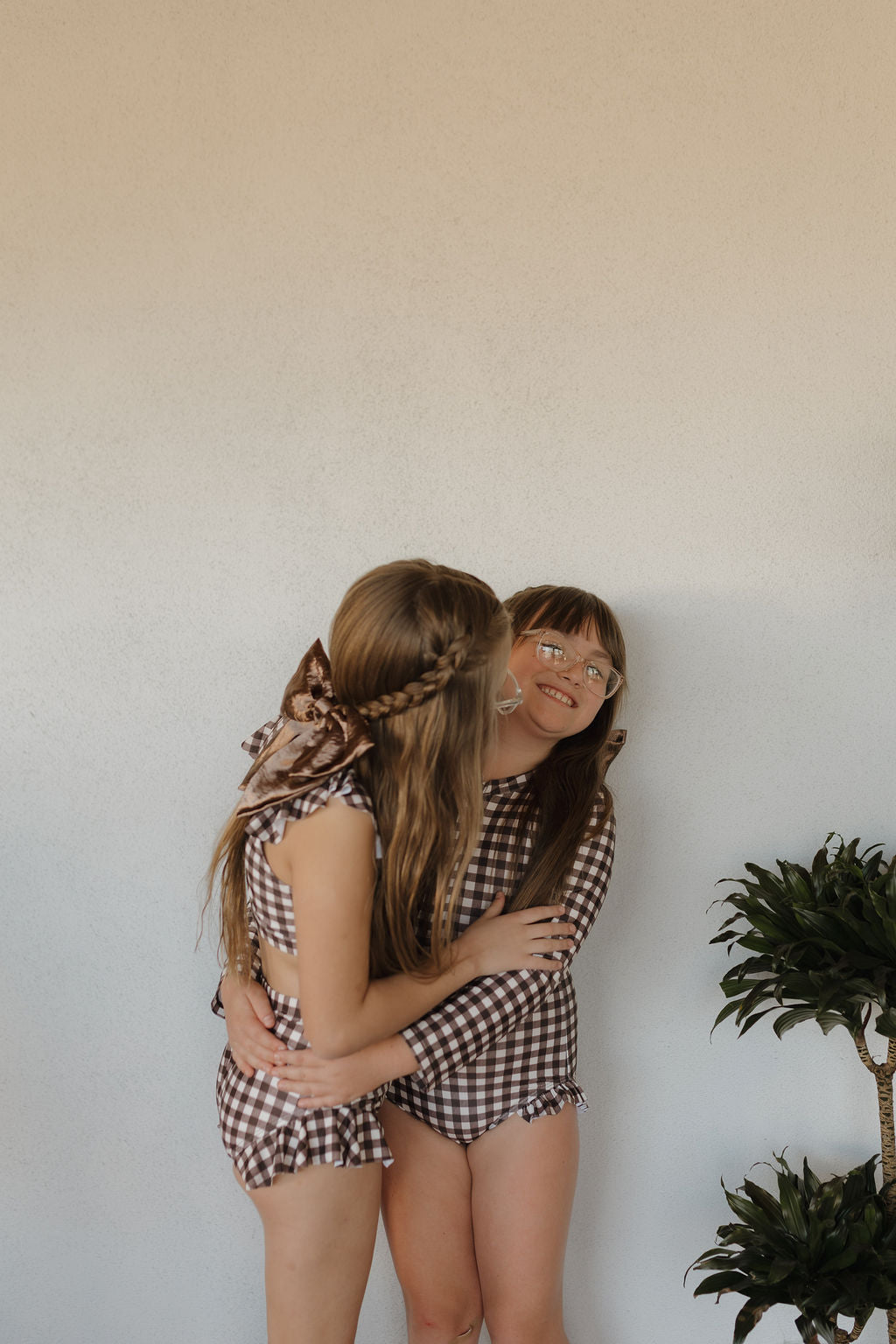 Two young girls from forever French baby joyfully pose in matching Girls Long Sleeve Swimsuits in Chocolate Gingham against a pale wall. One wears a bow and hugs the other, who smiles while wearing glasses. A small plant peeks in at the bottom right.