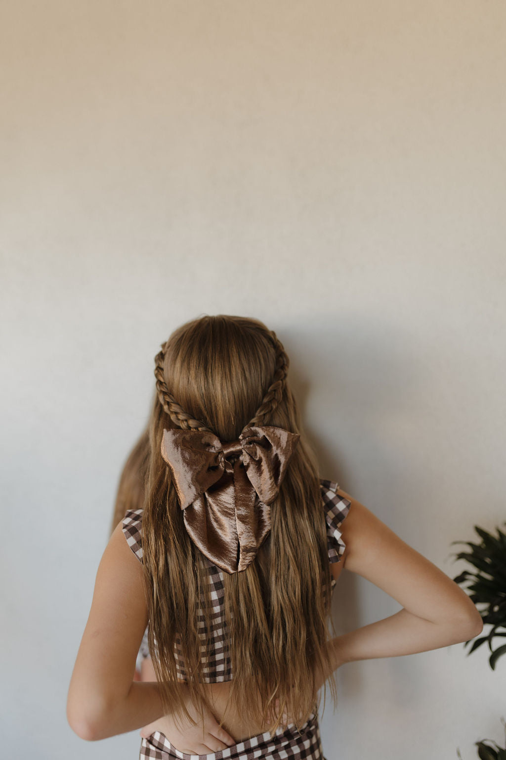 A person with long, wavy hair faces away, featuring a braid wrapped around the top of their head and a large brown bow. They're wearing forever french baby's Girls Two Piece Swimsuit from the Chocolate Gingham Collection, with a plant visible in the bottom right corner.