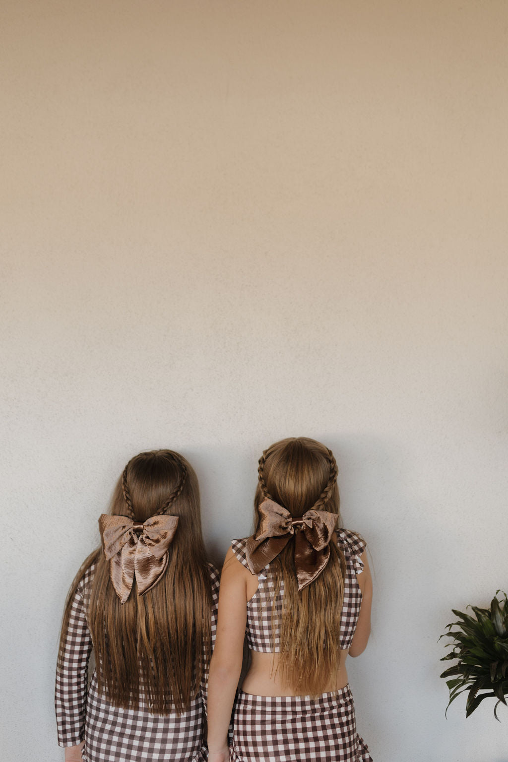 Two girls with long braided hair, wearing the Girls Long Sleeve Swimsuit from the Chocolate Gingham Collection by Forever French Baby, face a wall. Velvet bows adorn their outfits as a plant peeks against an off-white backdrop, completing this matching scene.