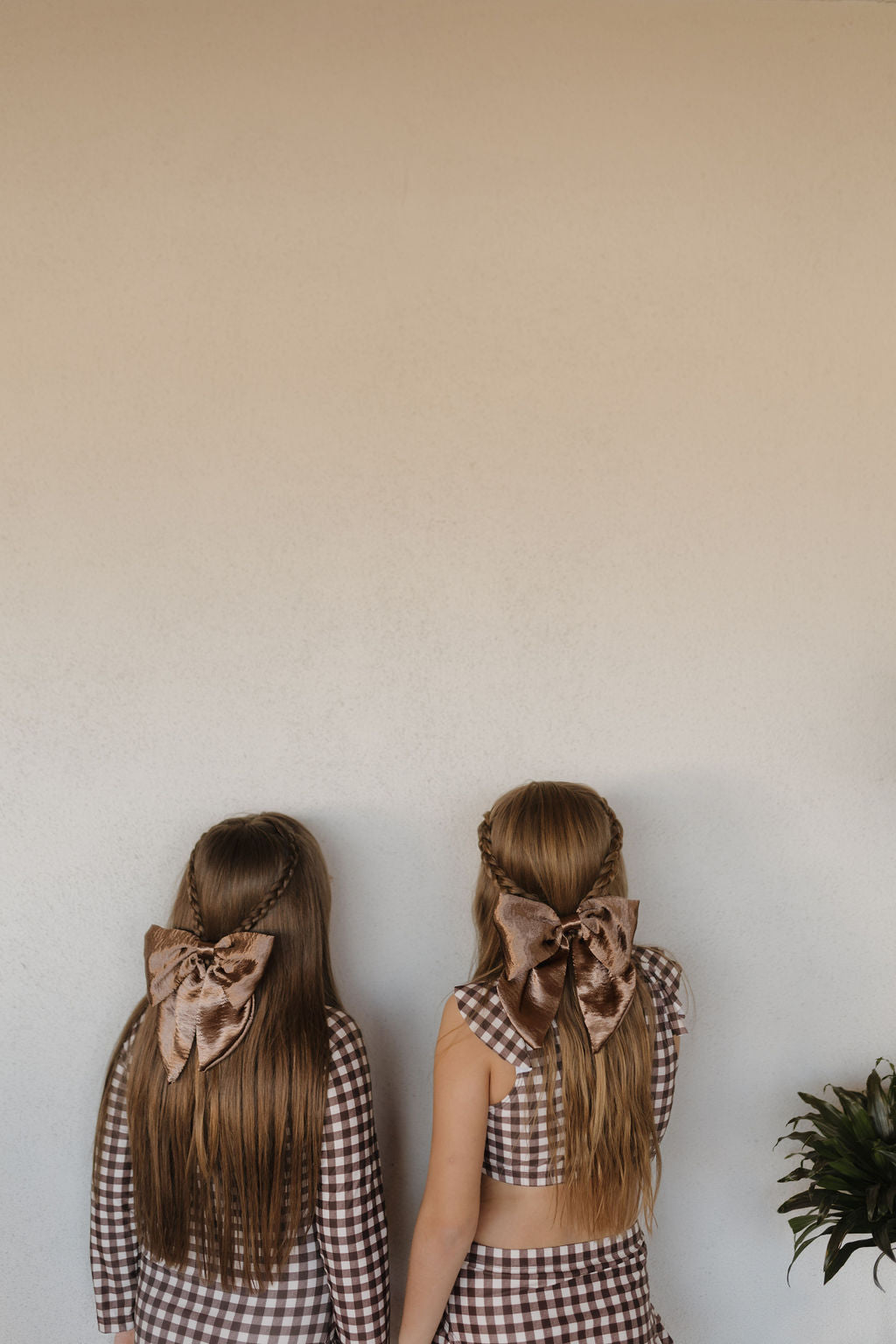 Two girls with long hair and large brown bows gaze at a beige wall, wearing the Girls Long Sleeve Swimsuit from the Chocolate Gingham collection by forever French baby. Their elegant look is enhanced by a partially visible plant in the bottom right corner.